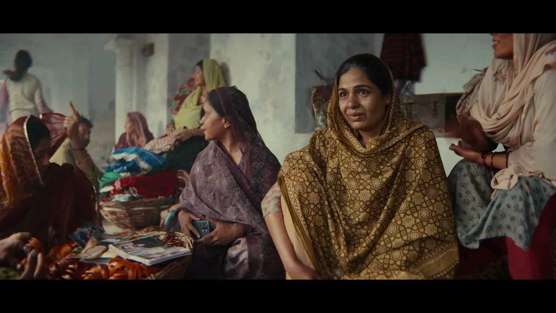 A group of women in traditional Indian attire are gathered in a room, some looking at items in baskets, others conversing.