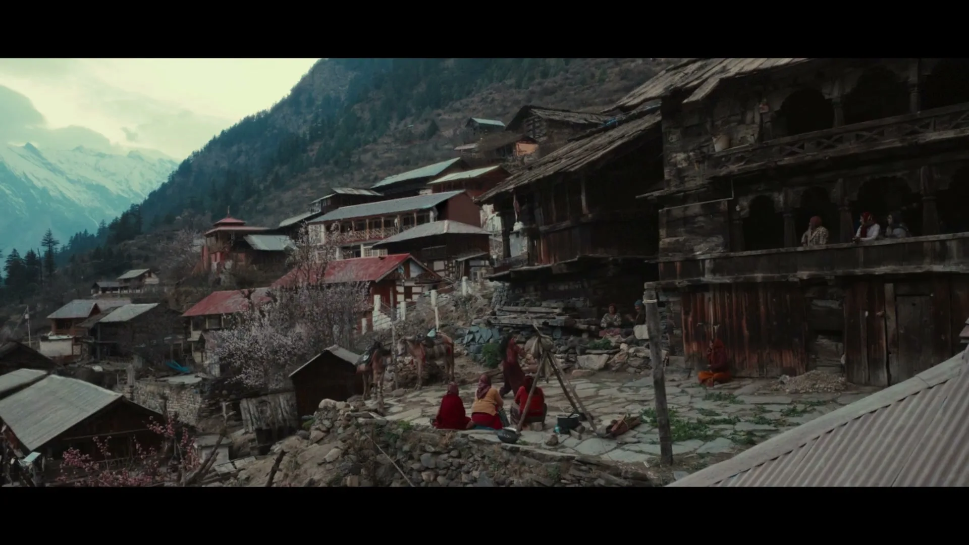 A wide shot of a mountain village with traditional wooden houses, showing people in traditional attire gathered in groups, with snow-capped mountains in the background.