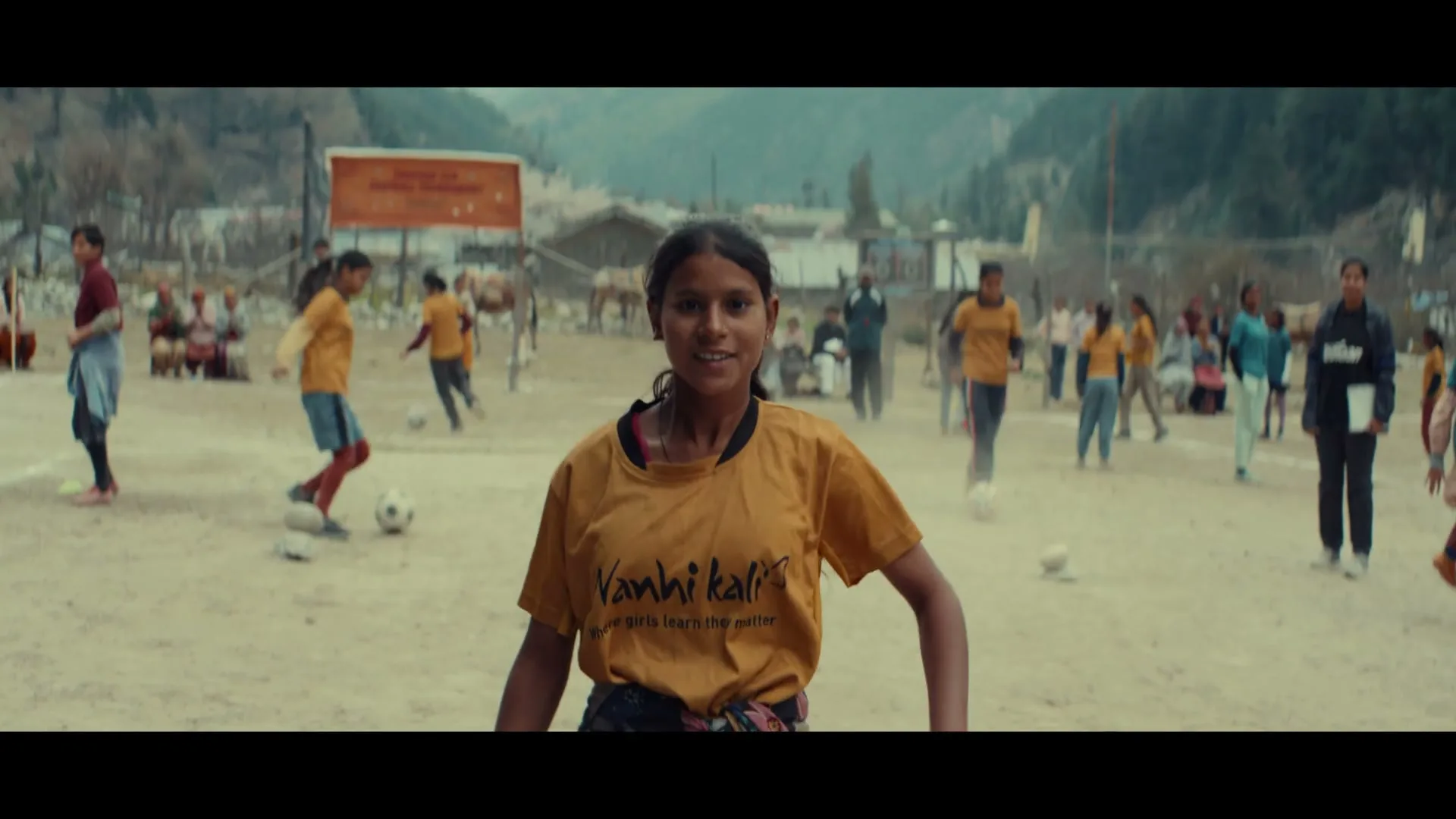 A young girl in a yellow t-shirt smiles at the camera while other girls play soccer in a dusty field with mountains in the background.