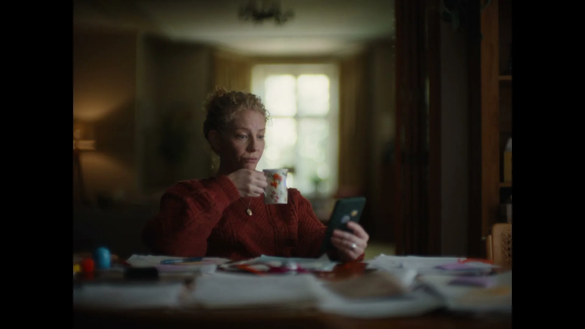A woman with red hair in a red sweater drinks from a mug and looks at her phone, then looks up thoughtfully at a window in a cozy room.