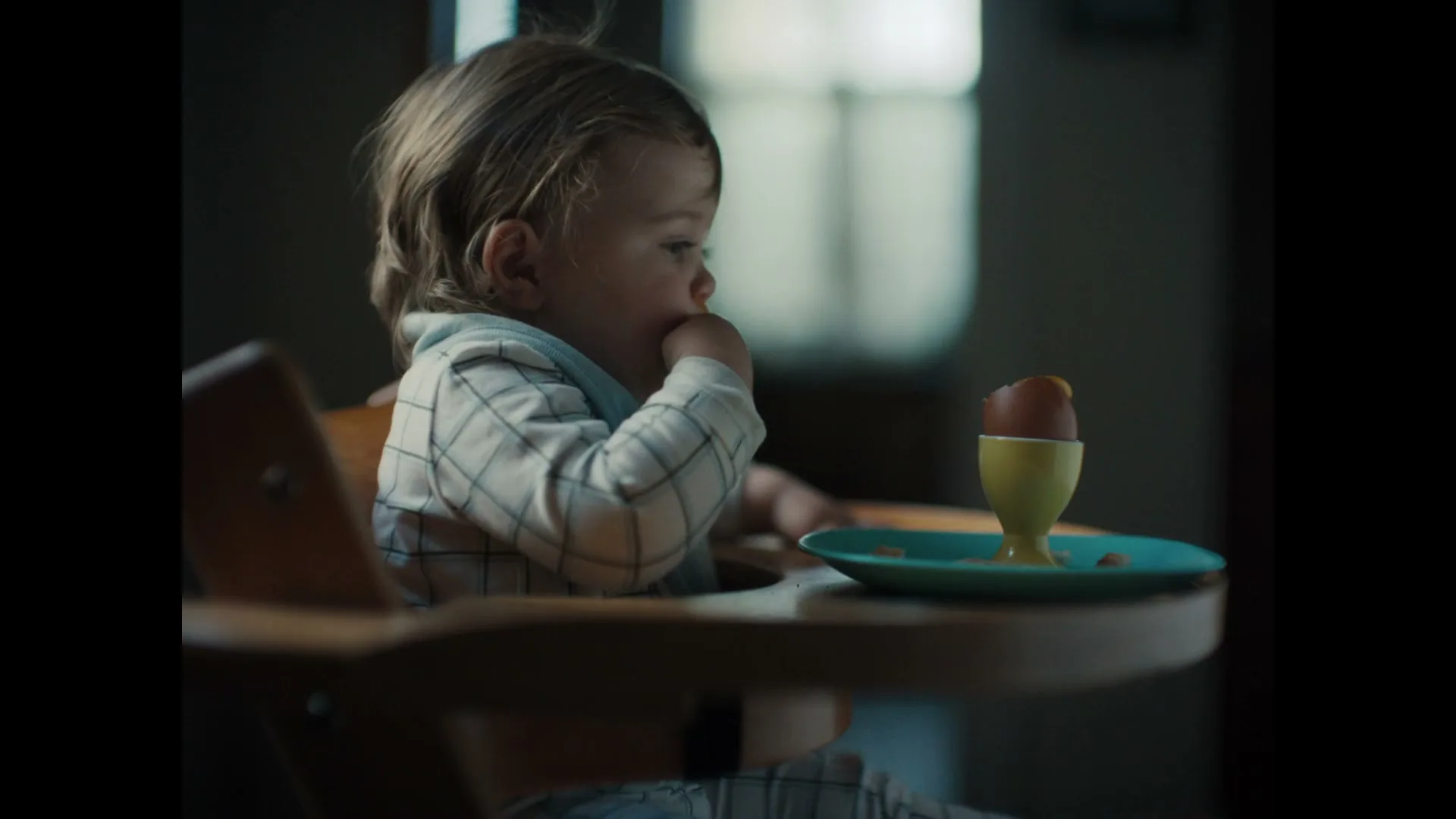 A baby with light brown hair in a high chair attempts to eat a soft-boiled egg from an egg cup on a blue plate in a dimly lit room.