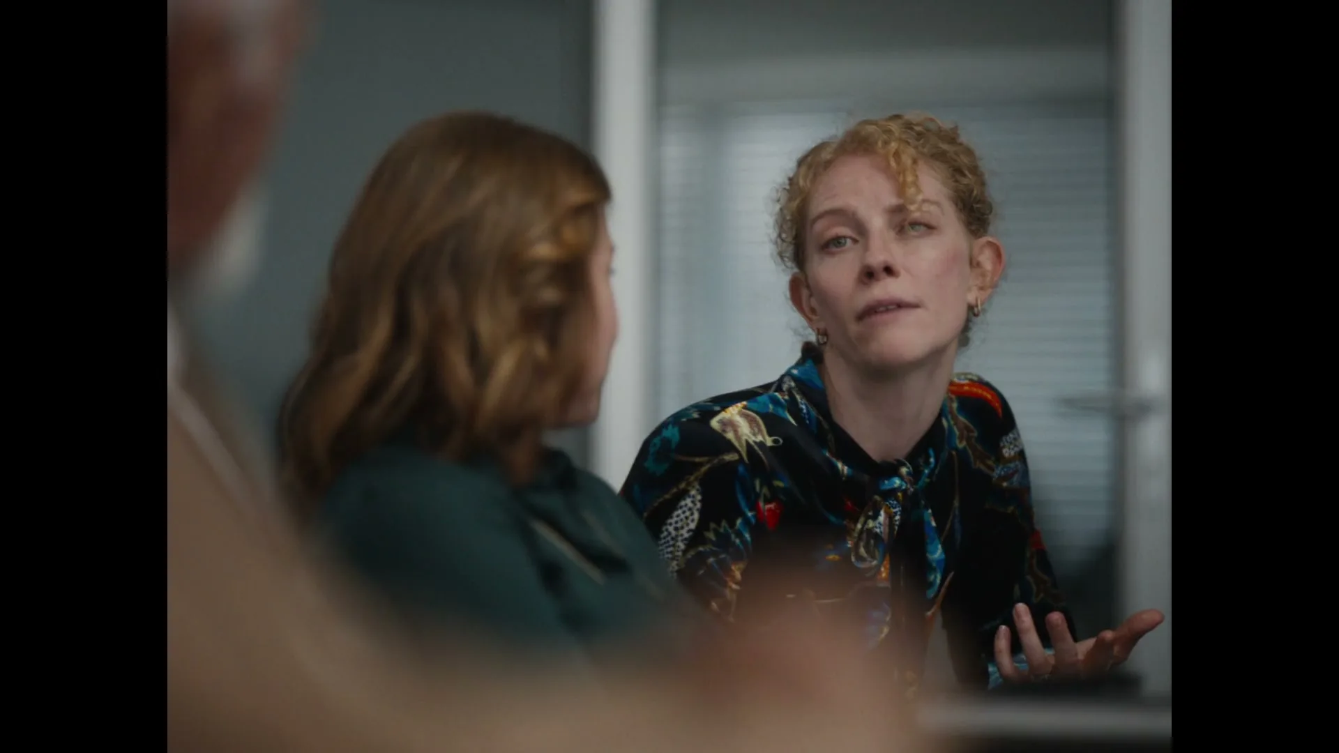 A woman with curly blonde hair in a colorful blouse talks animatedly, gesturing with her hands, while another woman with brown hair listens in an office setting.
