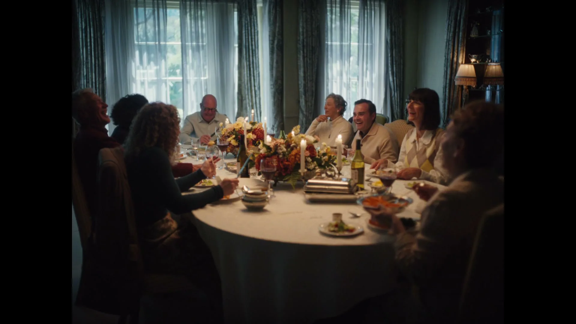 A diverse group of adults, including men and women, are gathered around a dining table, laughing and enjoying a meal together in a well-lit dining room.