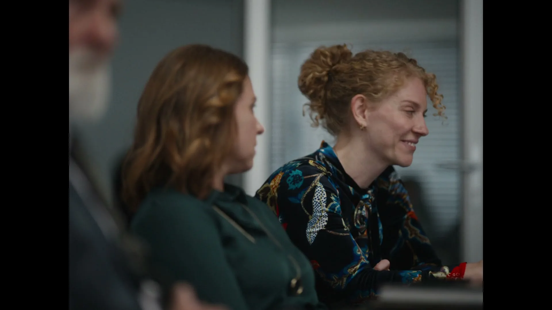 A woman with curly red hair in a patterned blouse smiles and looks at someone off-screen during a meeting in an office.