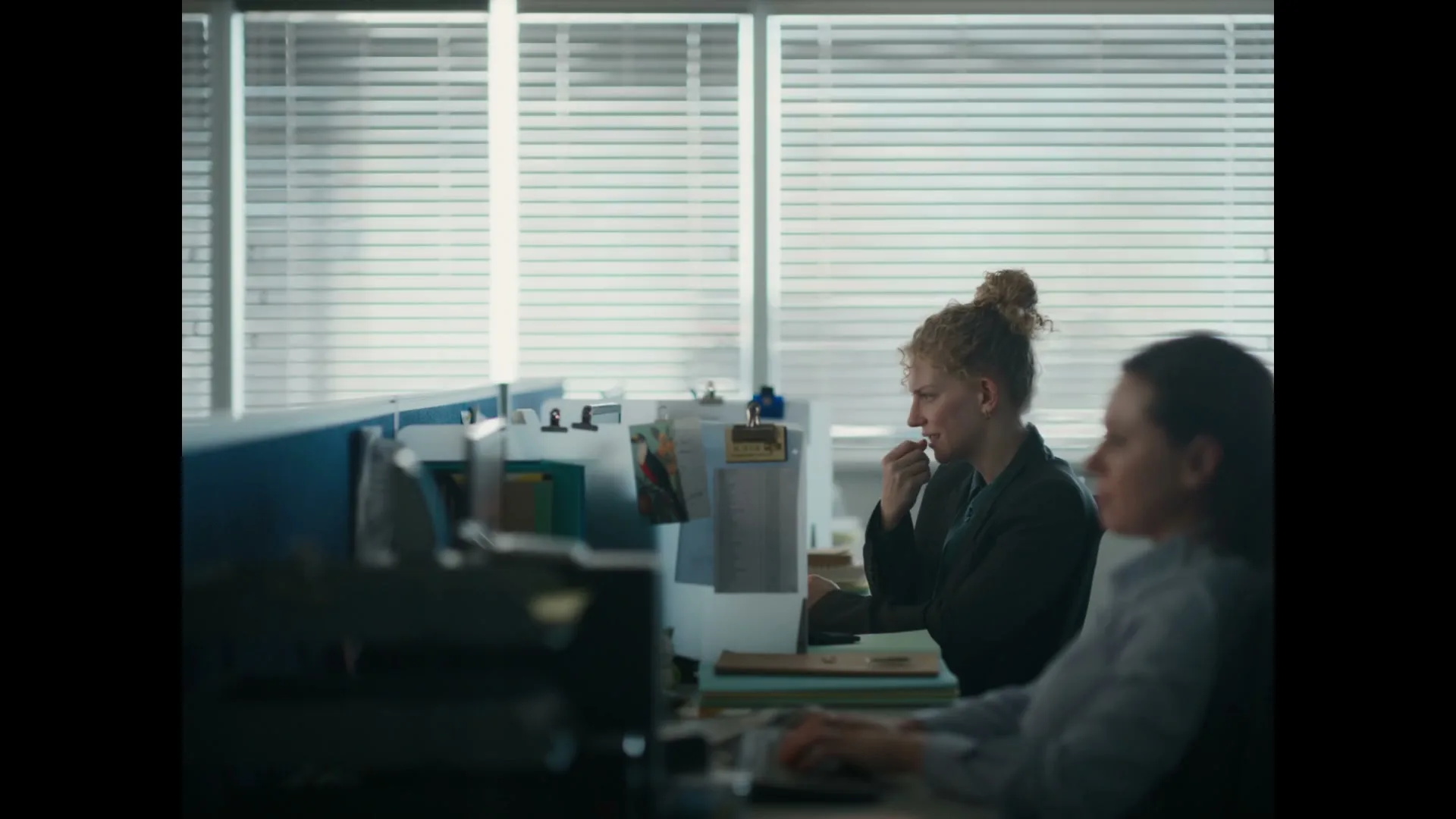 A woman with curly blonde hair in a bun works at a cubicle desk, looking at her computer screen in an office.