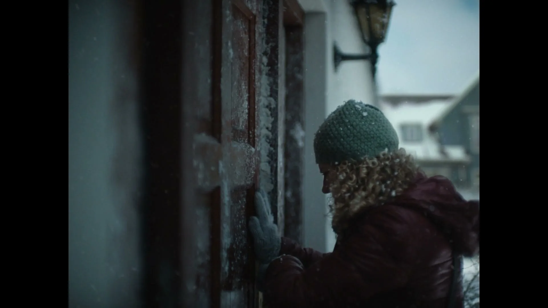 A young woman with curly hair and a green beanie struggles to open a snow-covered wooden door on a cold winter day.