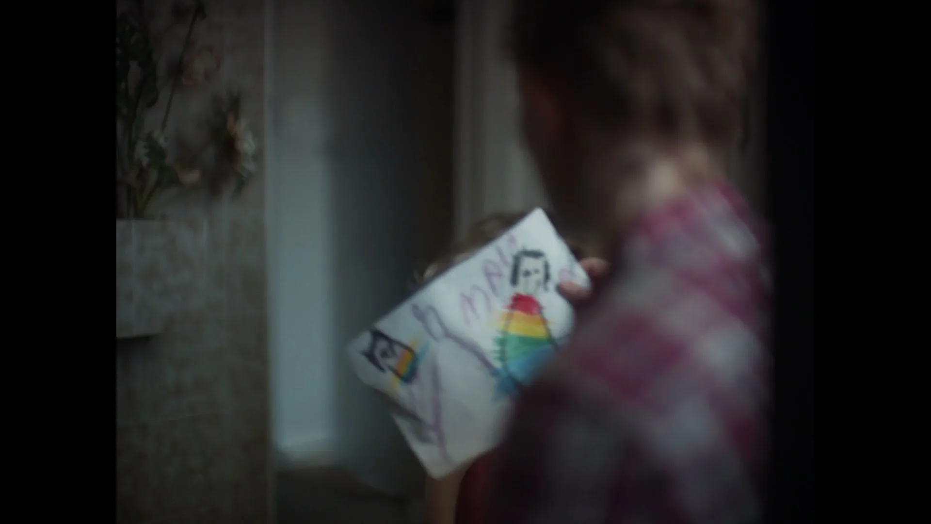A young child with curly hair excitedly shows a drawing to an adult, whose back is to the camera, in a dimly lit room.