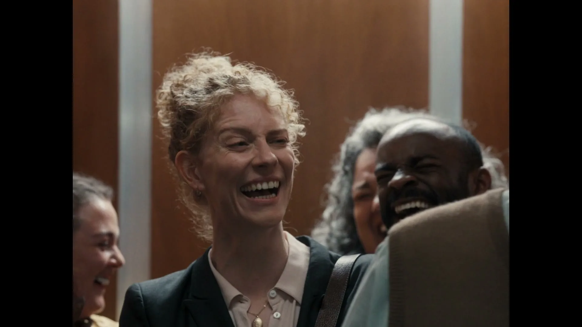 A diverse group of office workers, including a woman with curly blonde hair, laugh joyfully inside an elevator.