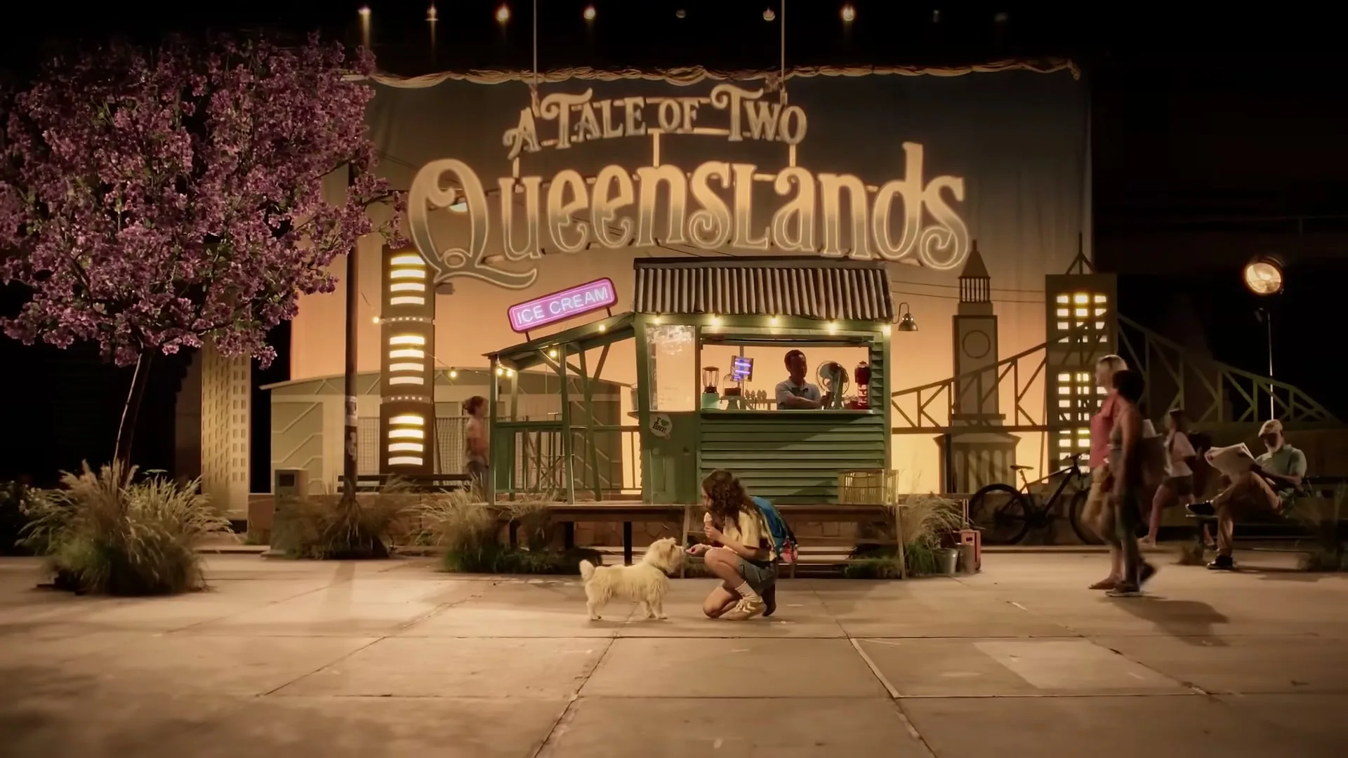 A young woman with long hair kneels to pet a small white dog in front of an ice cream stand and a large backdrop reading "A Tale of Two Queenslands" at night.