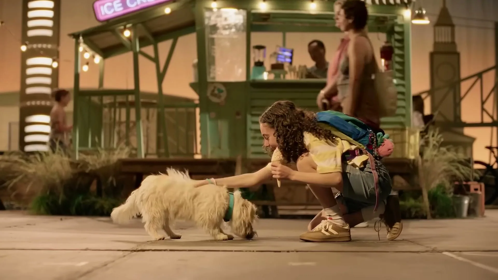 A young girl with curly hair, wearing a colorful backpack, crouches down to pet a small, fluffy white dog on a paved area in front of an ice cream stand at dusk.