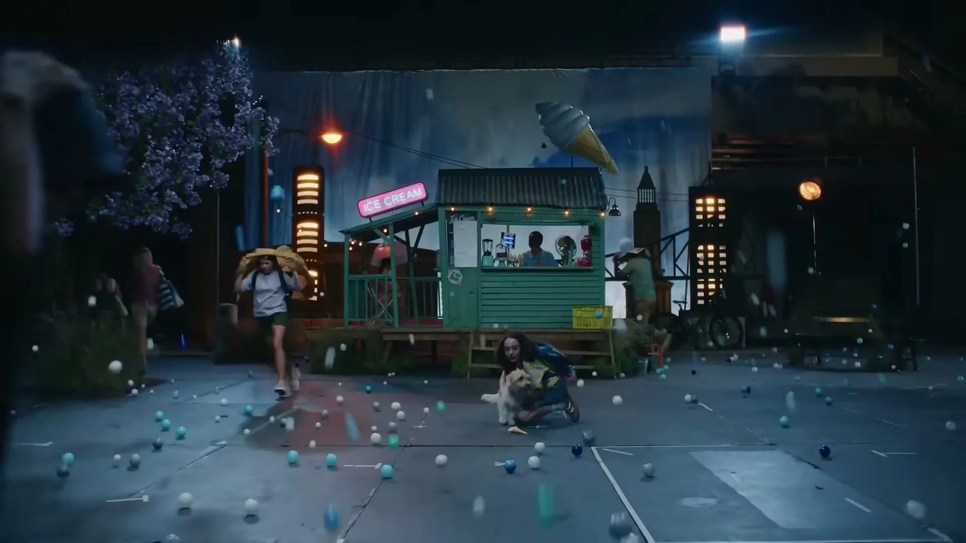 A young woman and her dog huddle on the ground as people run and ride bikes through a chaotic, windy outdoor set with an ice cream stand and falling balls.