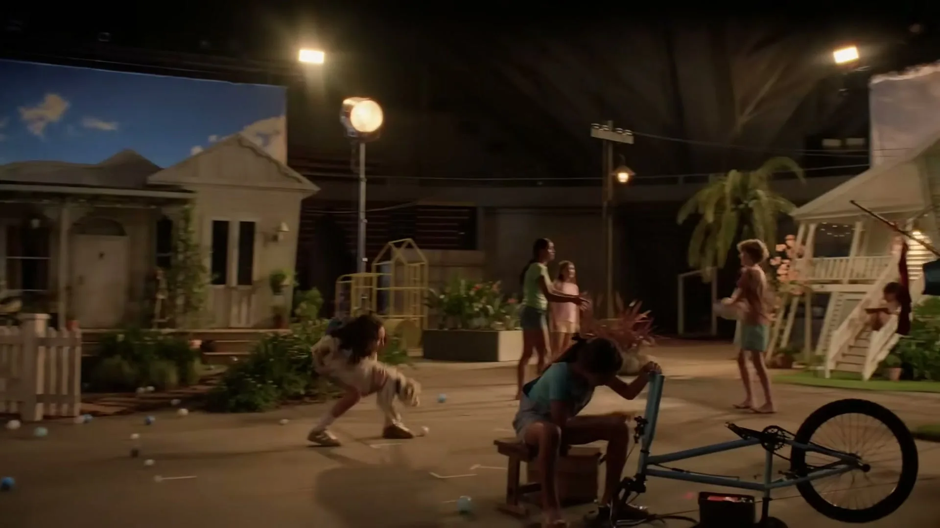 A group of children and a dog play in a street set on a soundstage, with houses and a sky backdrop.