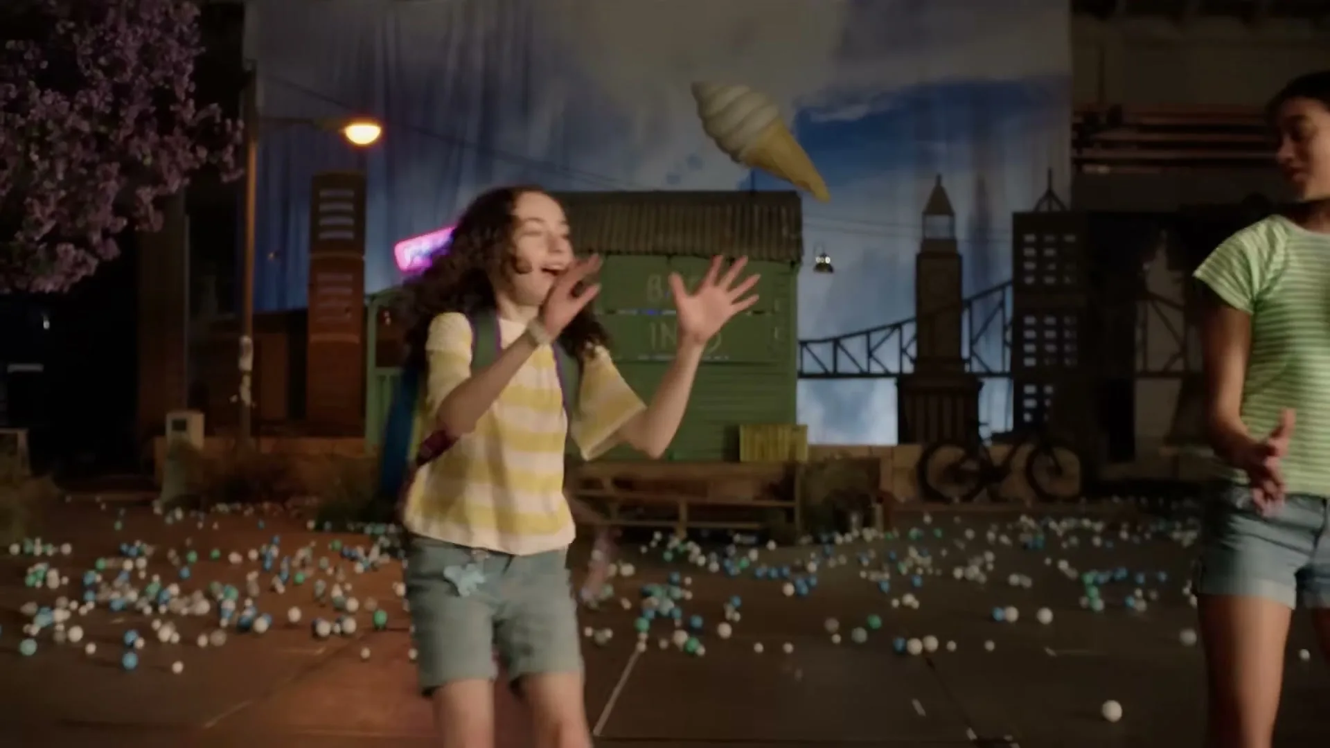 A young girl with curly hair and a backpack tries to catch a giant ice cream cone as it falls, surrounded by small balls on the ground in a stylized outdoor set.