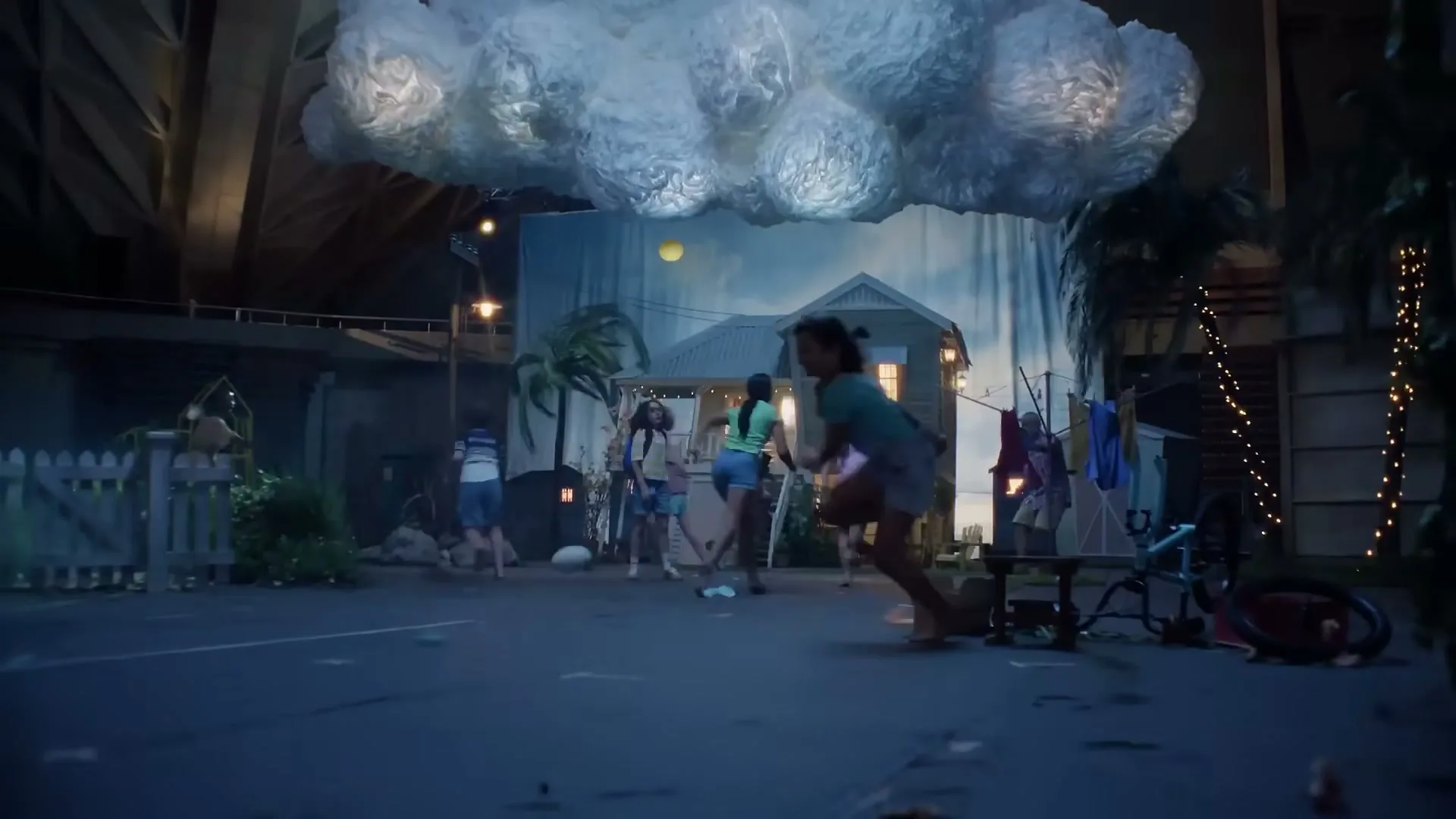 A group of children run and play in a stylized outdoor set, with a house backdrop and a large cloud prop overhead, as if a storm is coming.