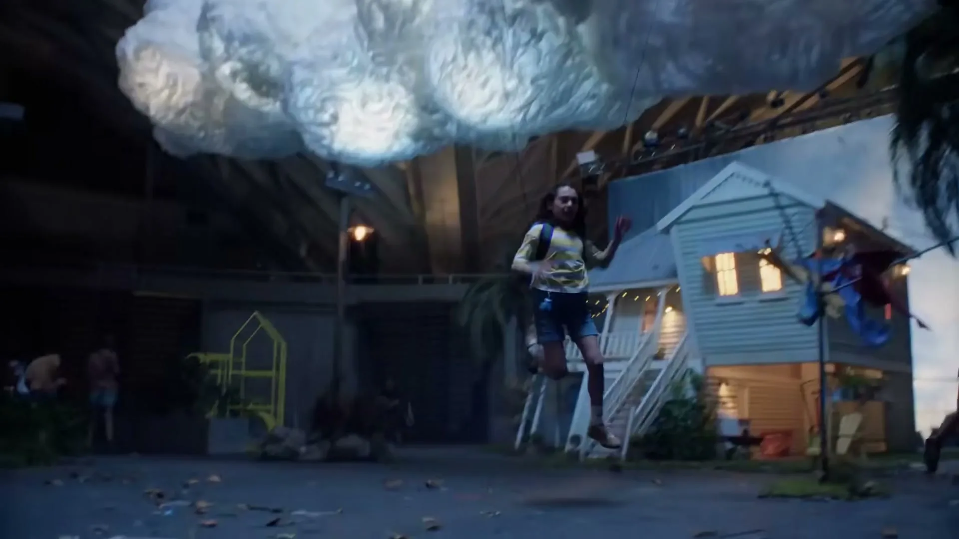 A young girl in a striped shirt and shorts runs across a set with a house and a large cloud prop, looking determined.