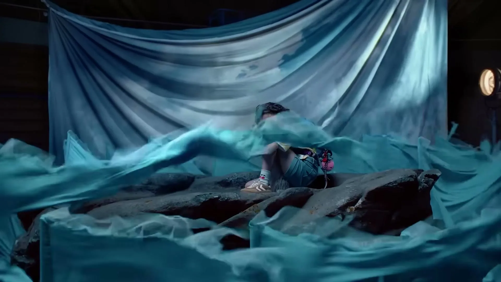A young person with long dark hair, wearing a colorful shirt and shorts, sits huddled on a rock as blue fabric waves around them in a studio.