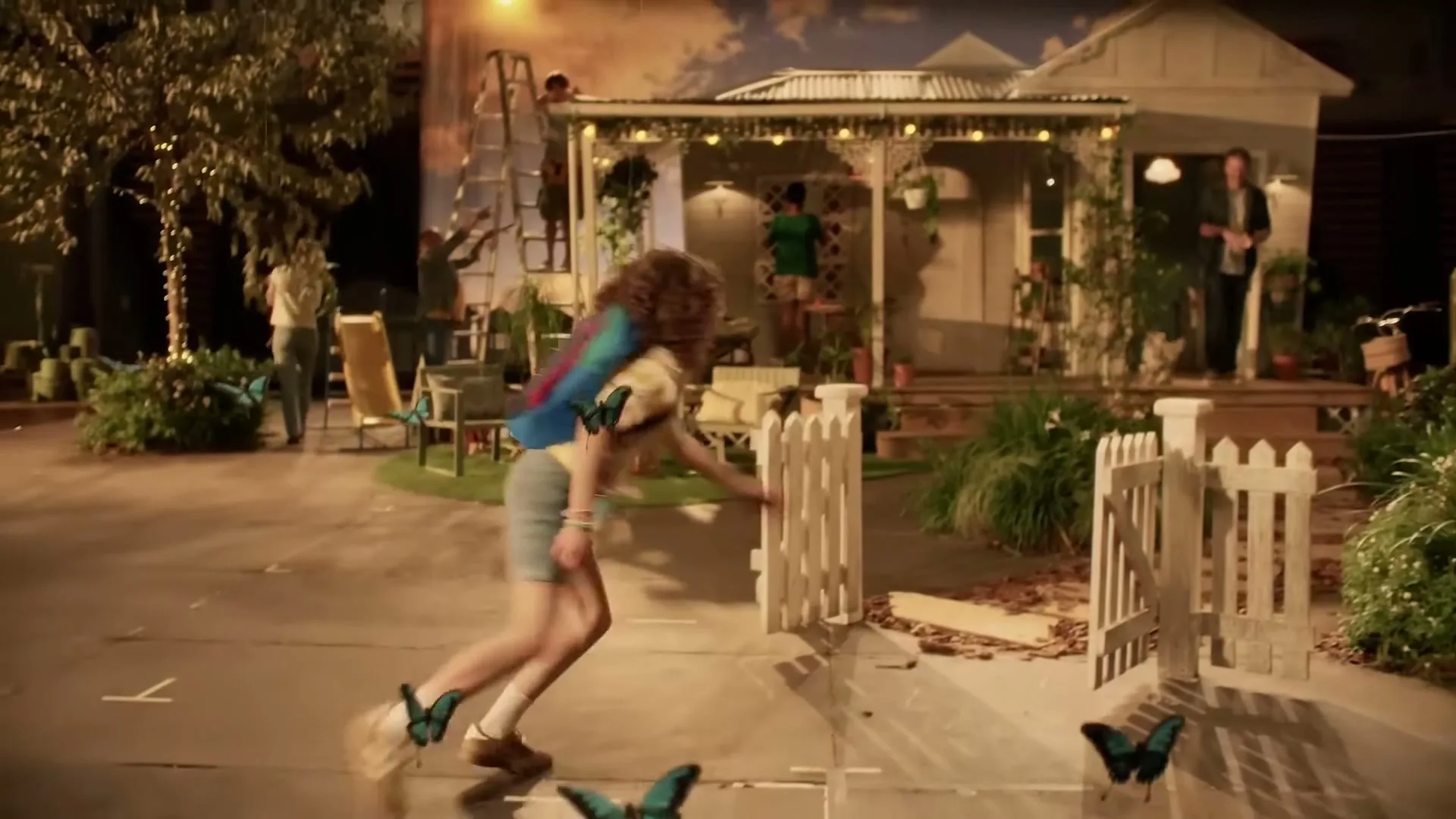 A young girl with a backpack runs towards a white picket fence, surrounded by blue butterflies, on a film set with a house facade in the background.