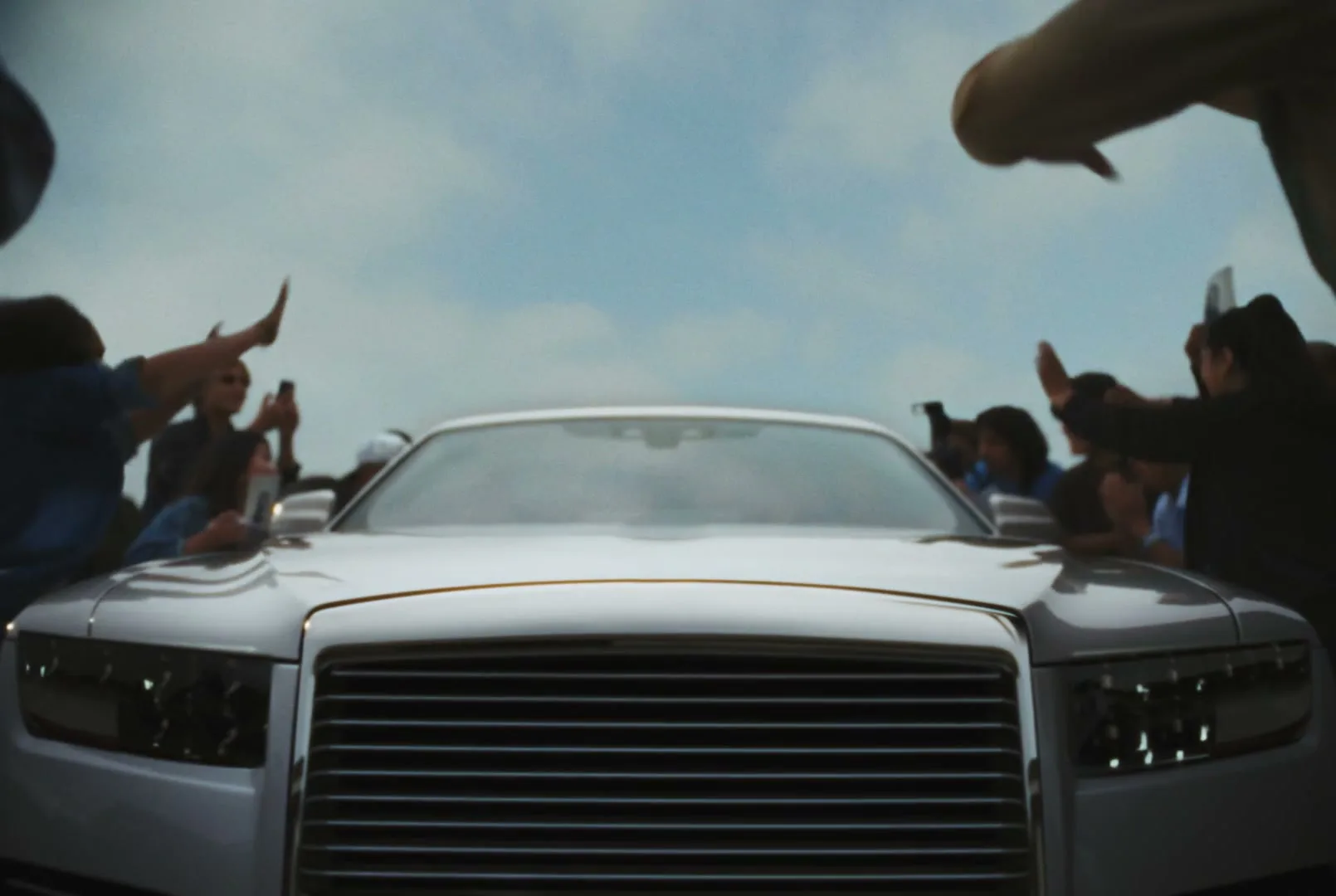 A white luxury car is surrounded by a crowd of fans with their hands raised, some holding phones, under a cloudy sky.