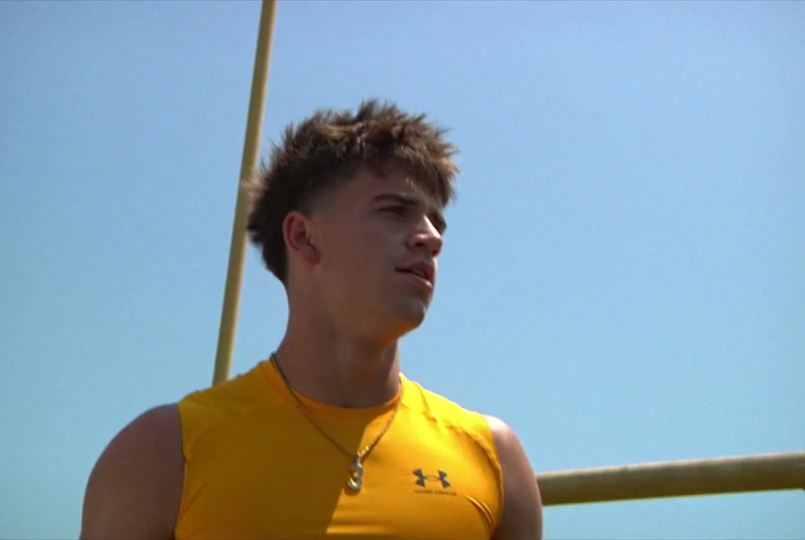 A young male athlete in a yellow shirt performs a backflip over a goalpost on a sunny day at a football field, then looks at the camera and claps.