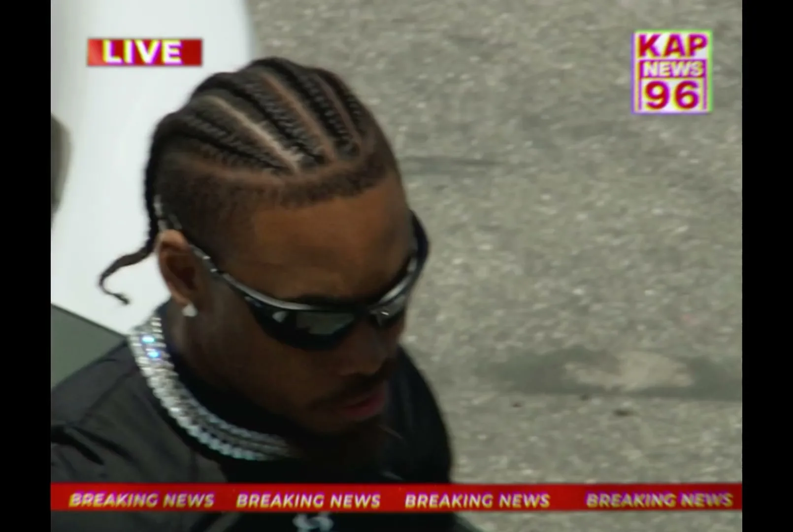 A male football player with braids and sunglasses exits a white car, wearing a black t-shirt and diamond chains, as news graphics overlay the scene.