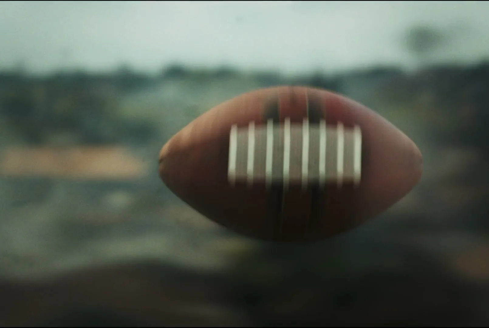 An American football spins through the air, blurred against a hazy, out-of-focus background of an outdoor landscape.