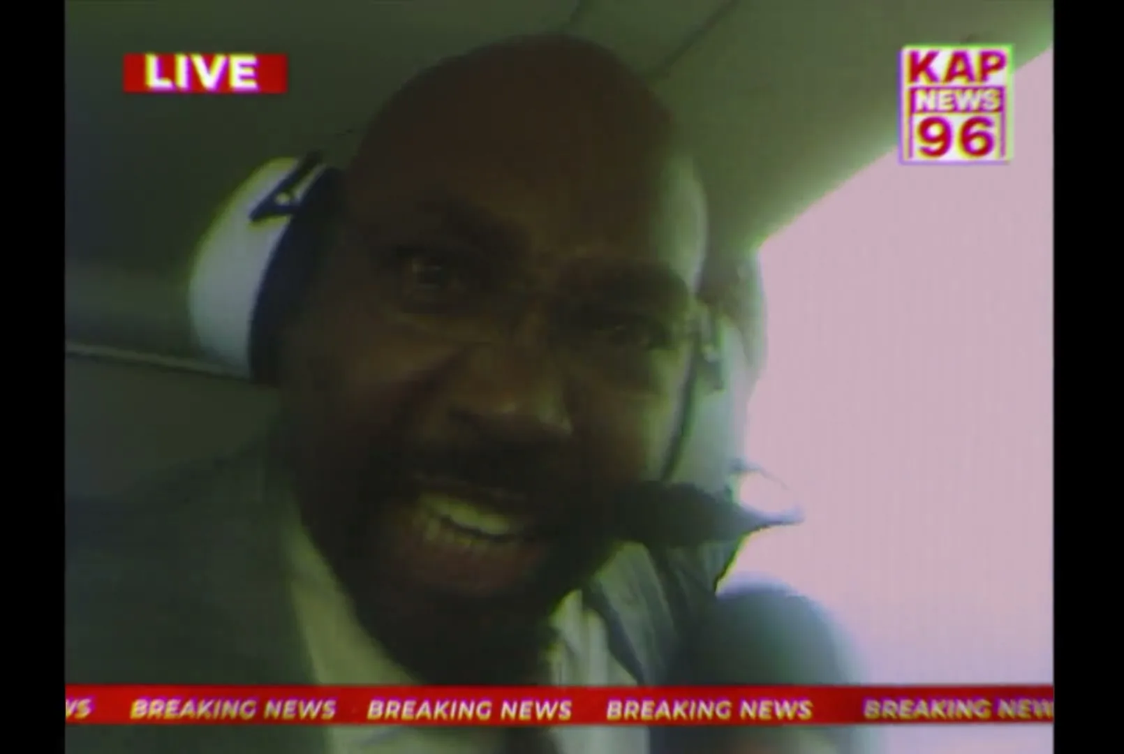 A bald African-American man in his 40s, wearing a suit and headphones, shouts into a microphone from inside a helicopter, with a news channel overlay.