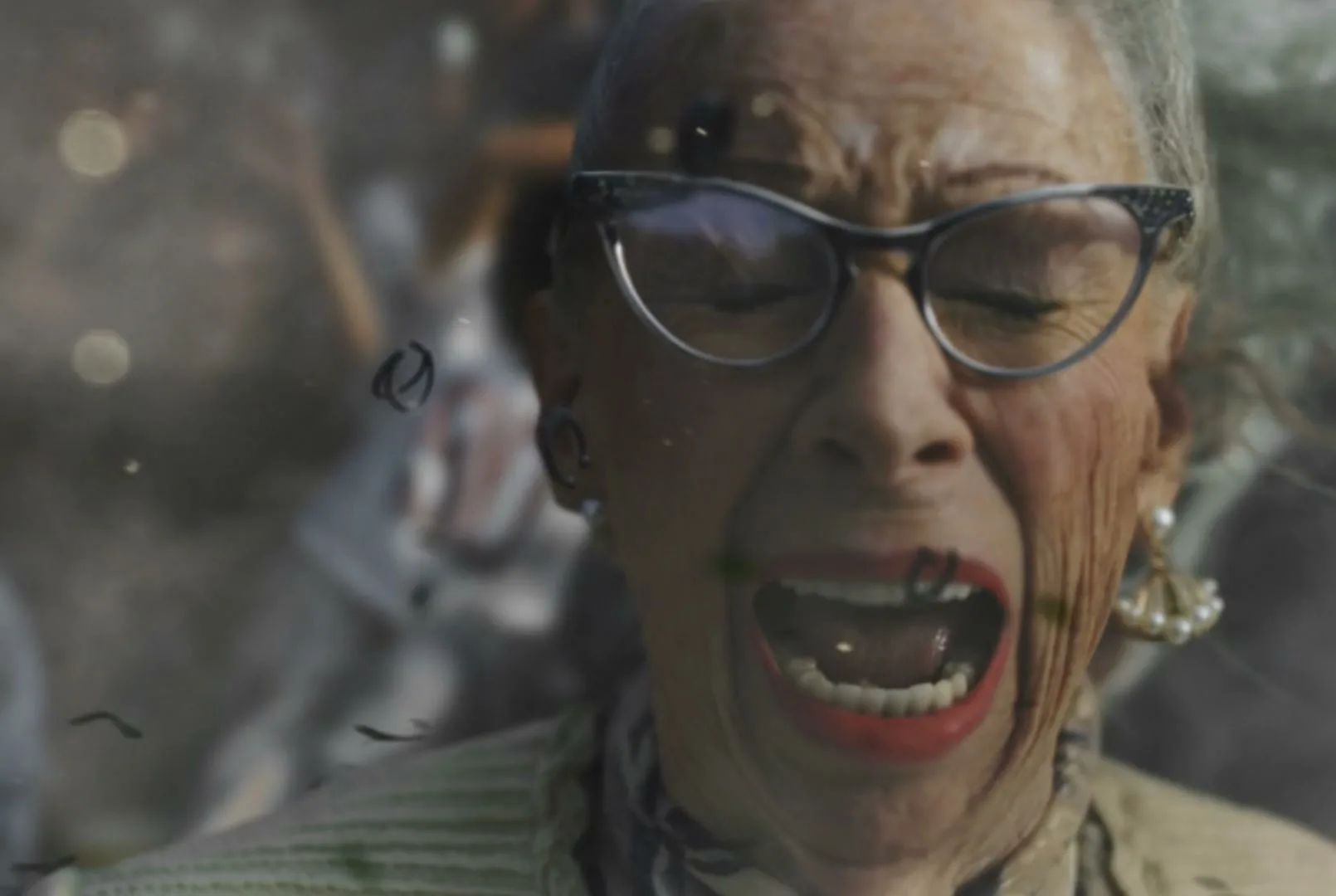 An elderly woman with glasses and red lipstick screams in terror as debris flies around her, possibly from an explosion or impact.