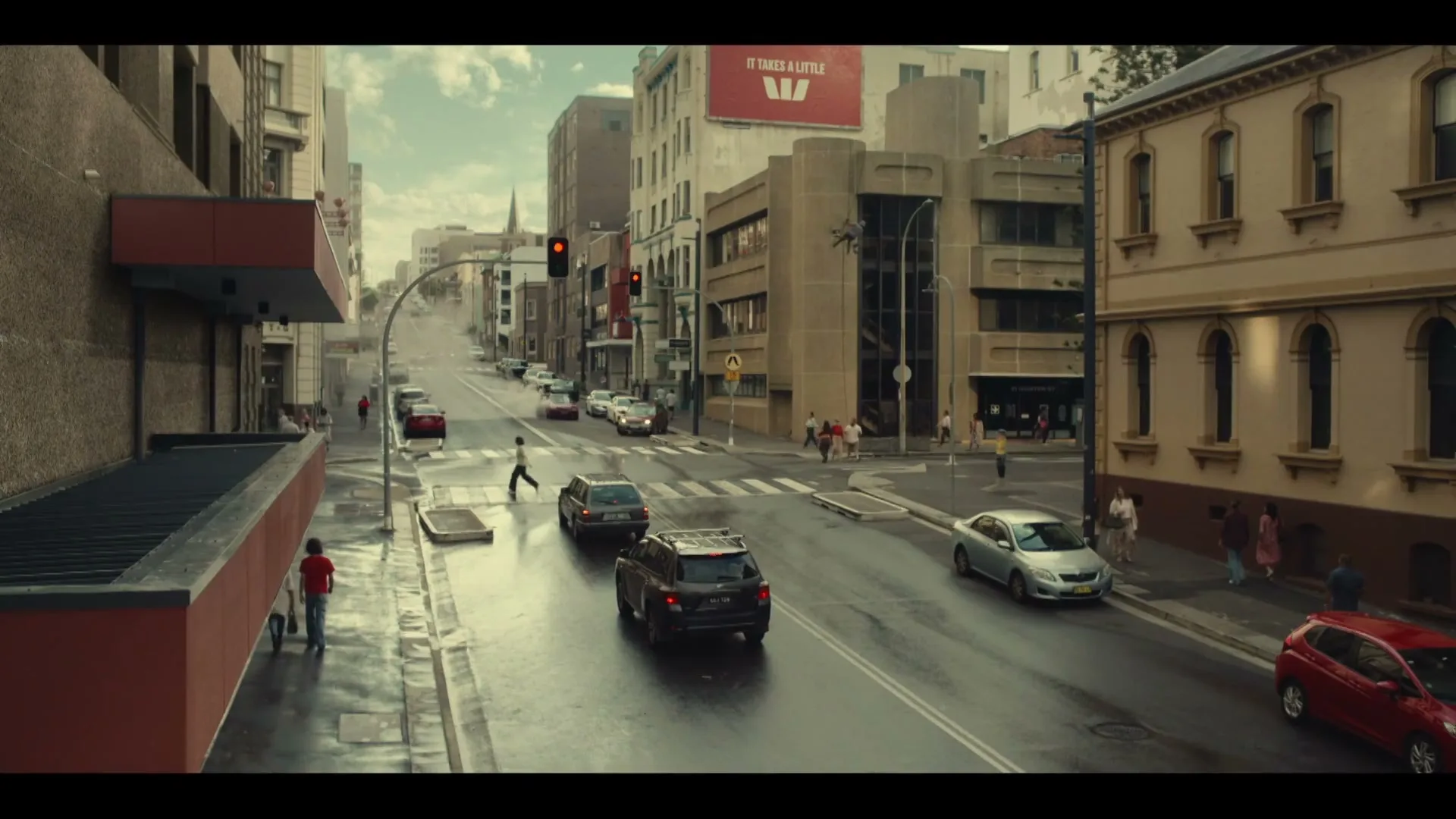 A wide shot of a city street with cars driving and people walking on the sidewalks, under a cloudy sky.