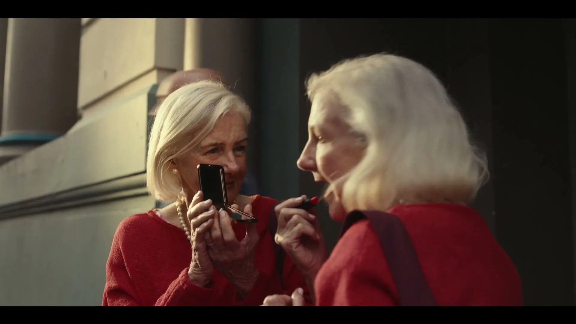An older woman in a red sweater applies lipstick while looking into a compact mirror held by her doppelganger on a city street.