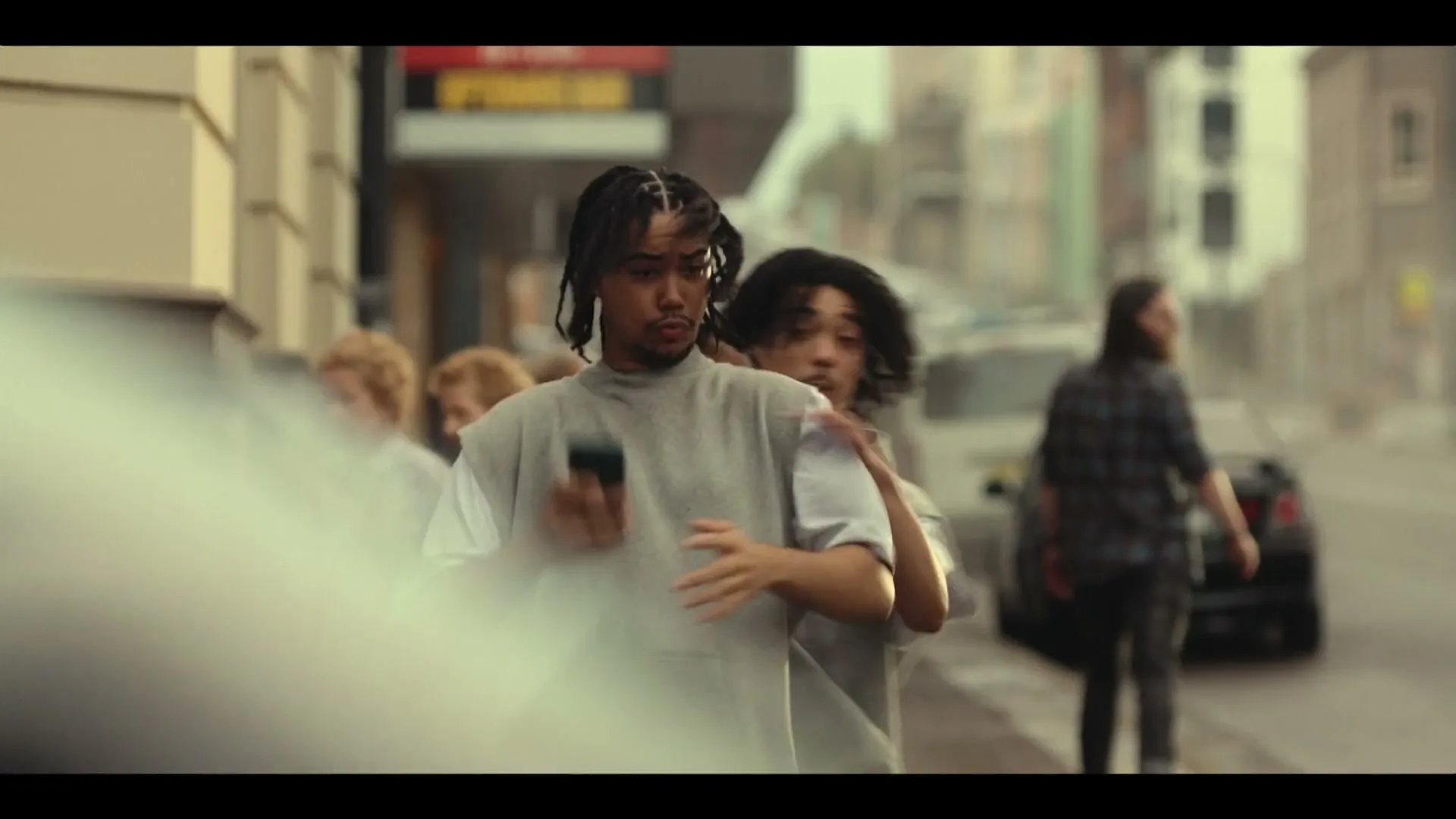 A young man with dreadlocks walks down a city street, looking at his phone, when another man identical to him bumps into him.