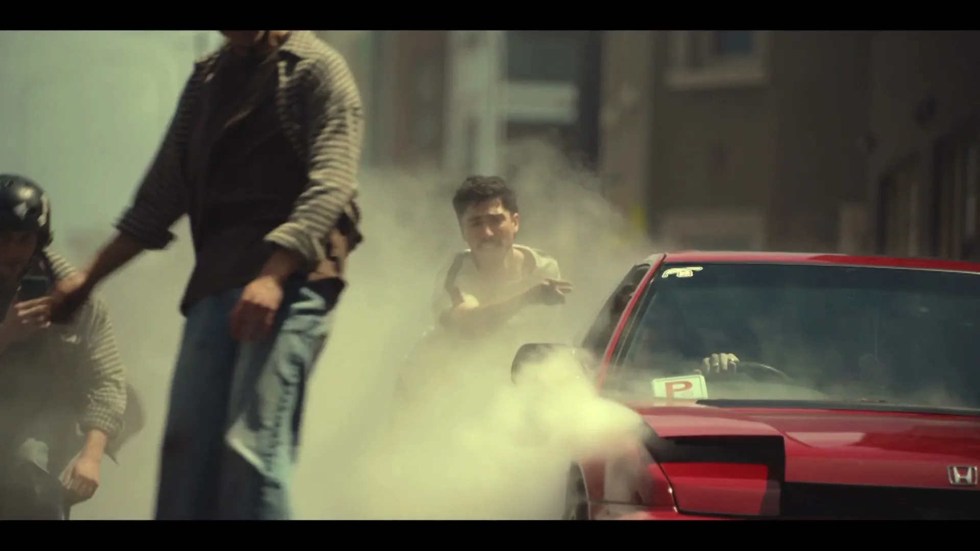 A group of young men are gathered around a red car doing a burnout, with smoke billowing around them on a city street.