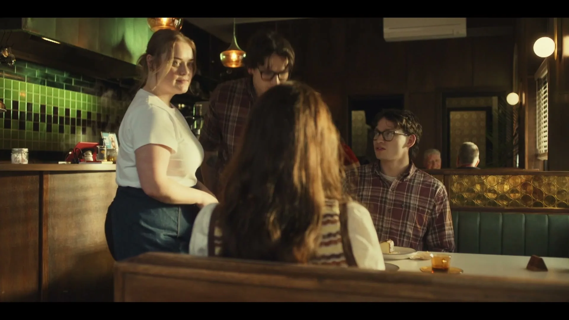 A waitress in a white t-shirt and blue skirt stands next to a booth where two young men in plaid shirts and a woman with long brown hair are seated in a dimly lit restaurant.