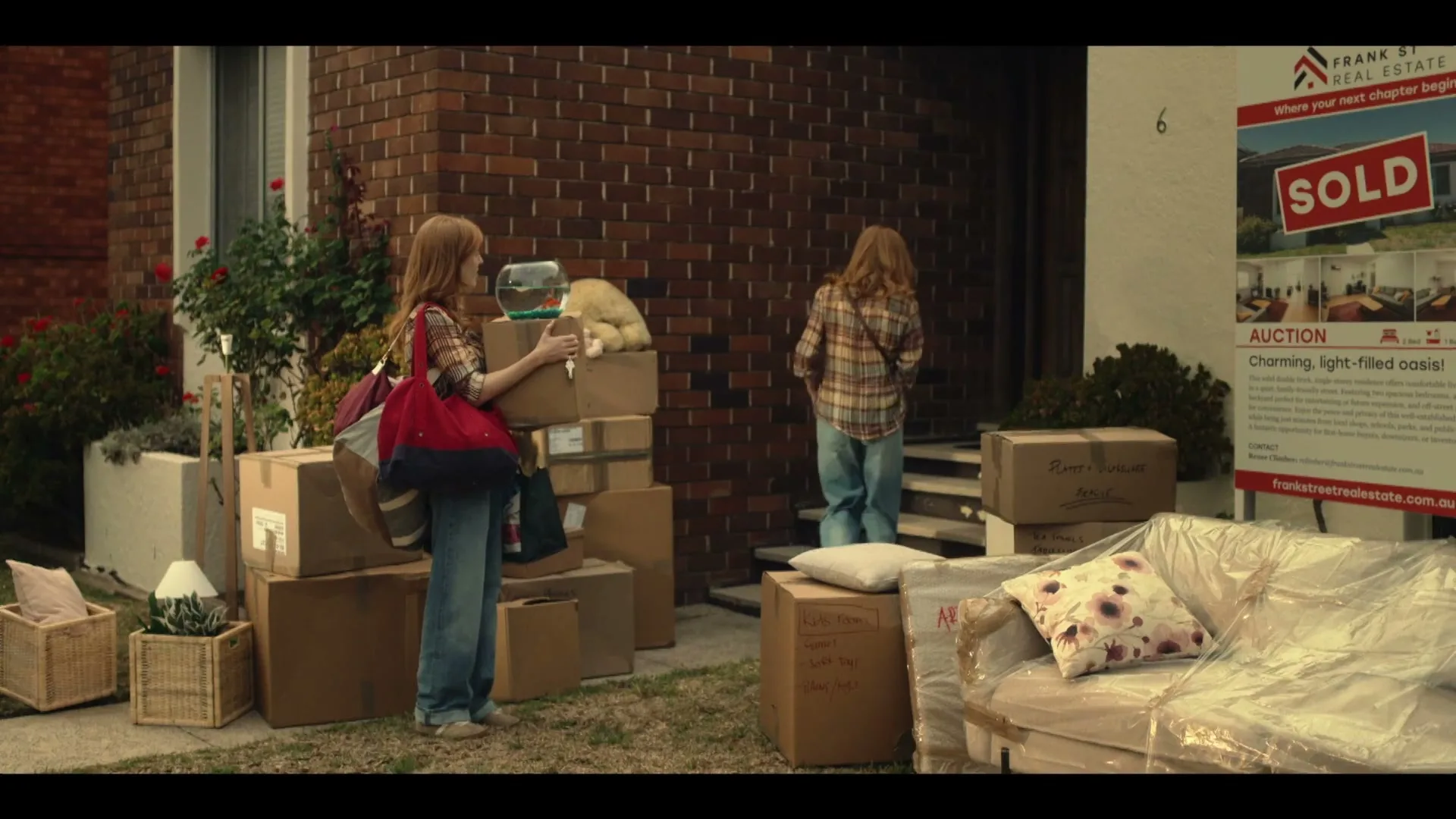 Two identical women with long red hair, wearing plaid shirts and jeans, move boxes and furniture into a brick house with a "SOLD" sign in the front yard.