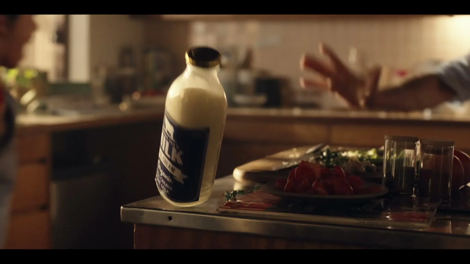 A bottle of milk and chopped tomatoes sit on a kitchen counter as a person's arm reaches out, in a warm, natural light setting.
