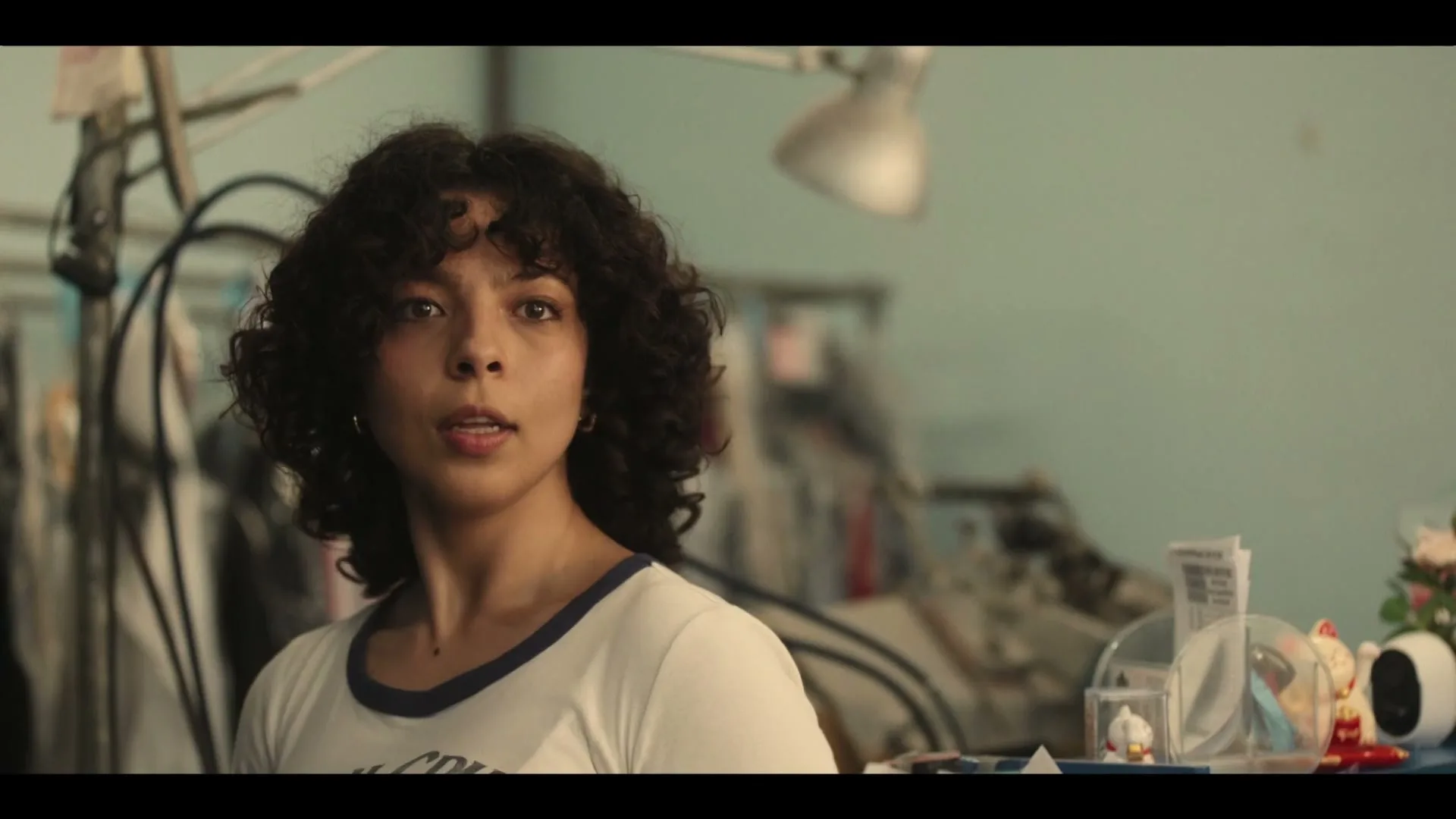 A young woman with curly hair, wearing a white t-shirt, looks surprised then smiles in a cluttered room with clothes racks and a desk lamp.