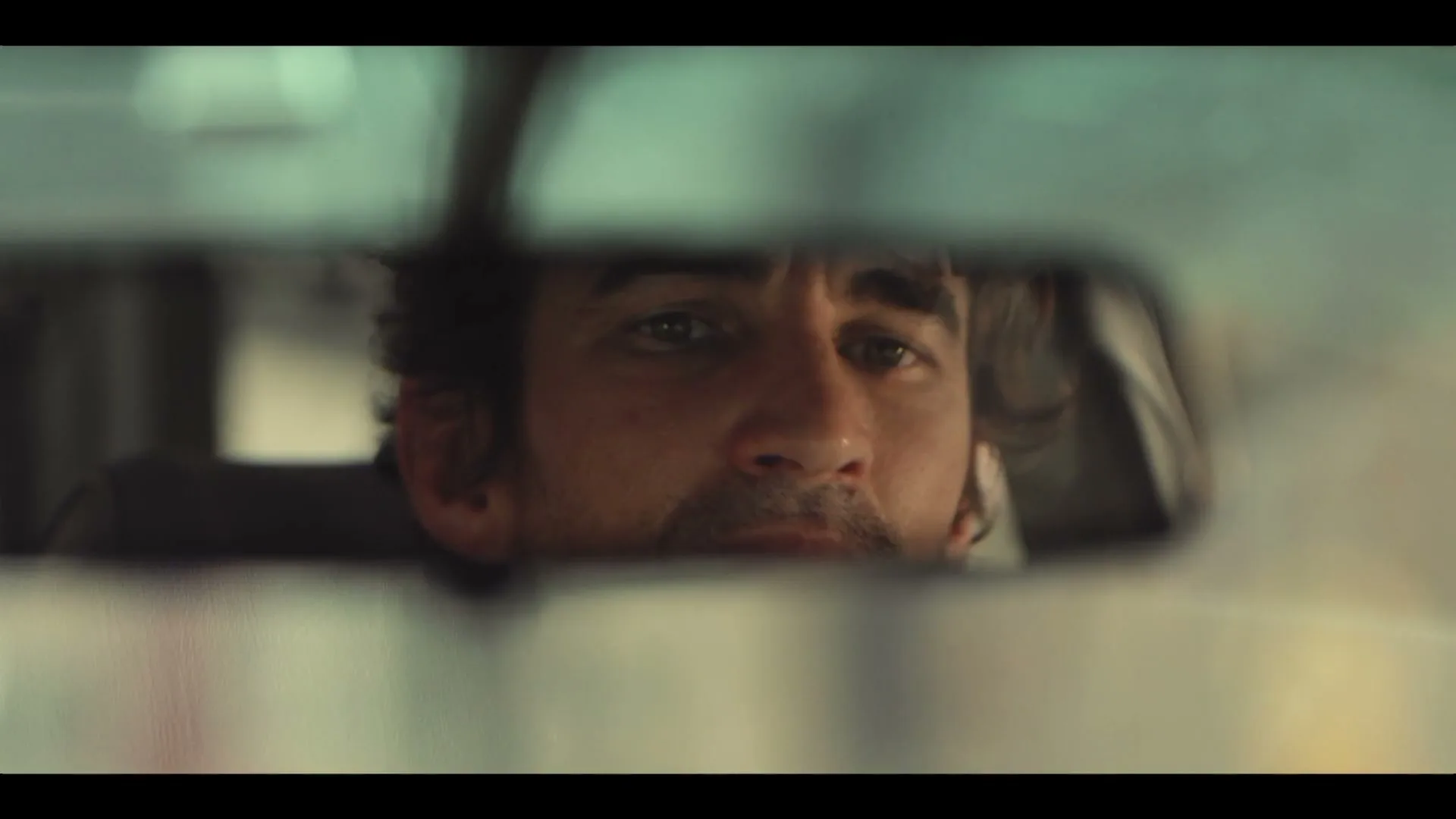 A man with dark, curly hair and a beard is reflected in a car's rearview mirror, looking forward with a neutral expression while driving.