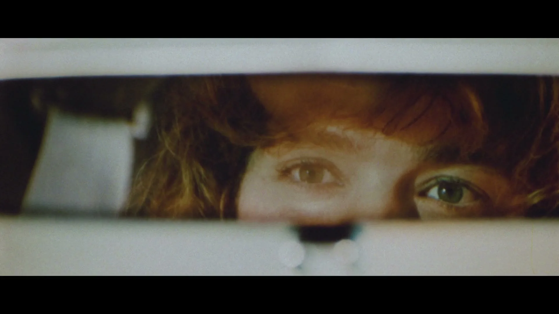 A woman with red hair and green eyes is seen in a car's rearview mirror, looking intently at something off-screen.