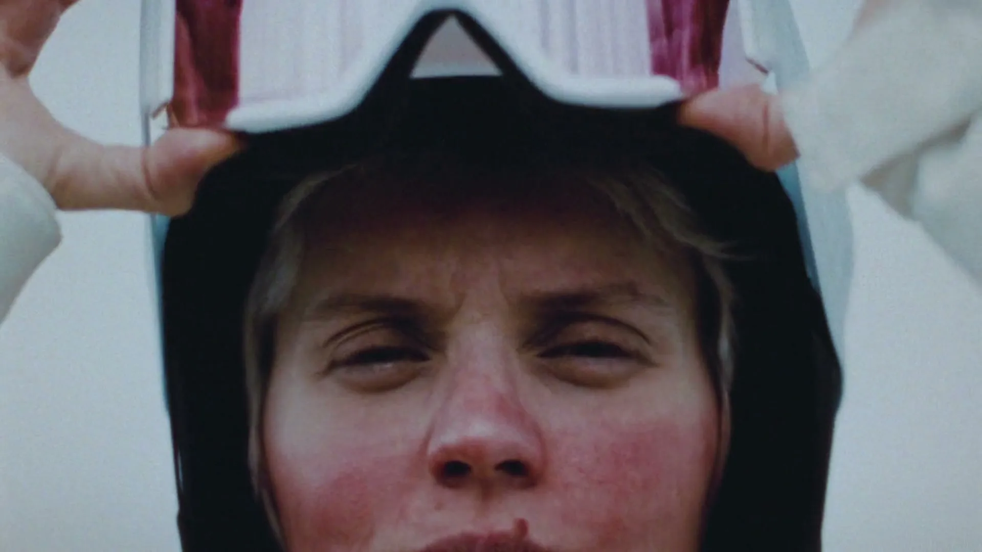 A woman with a red face and blonde hair takes off her ski goggles, squinting, in a close-up shot.