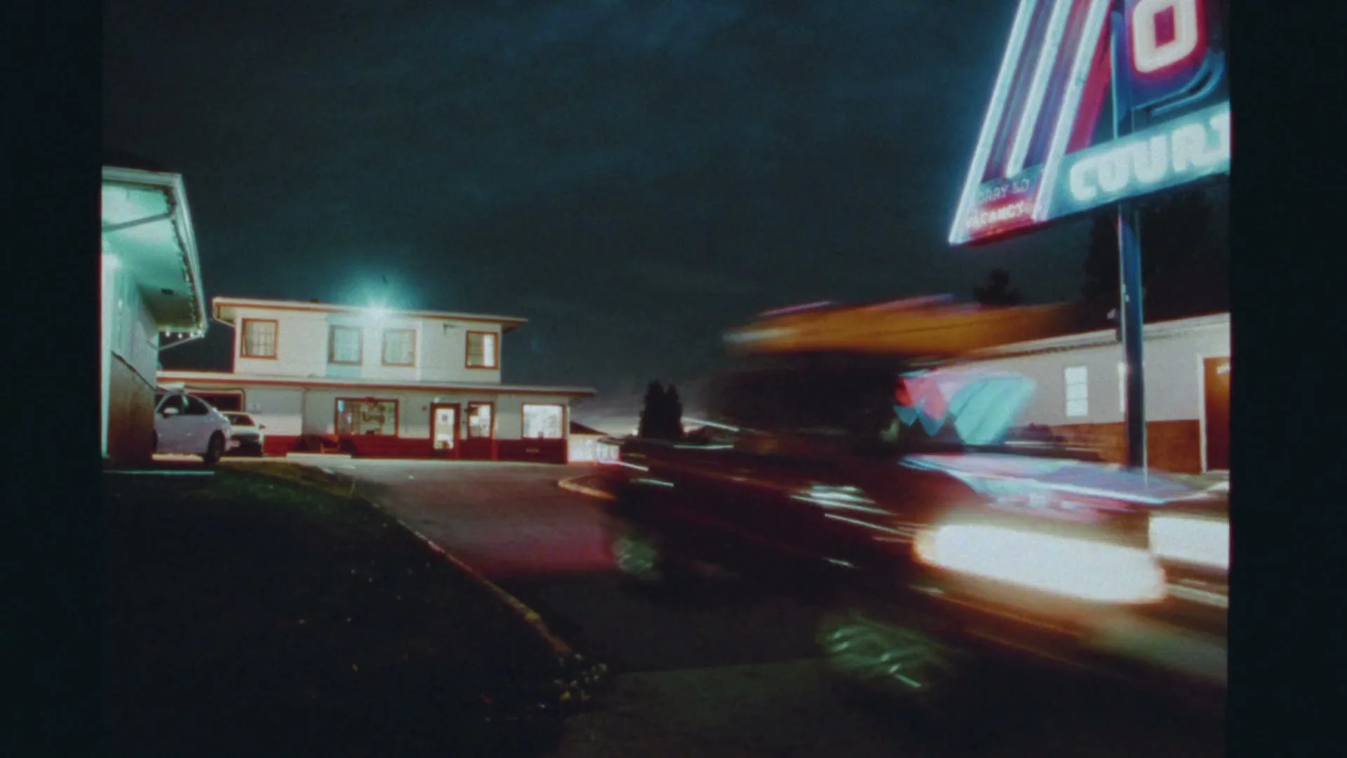 A red car with a surfboard on top speeds past a motel sign at night, leaving light trails.