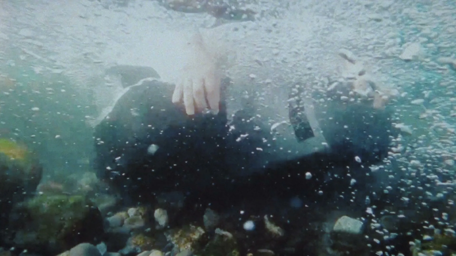A person in a dark wetsuit is seen underwater, surrounded by bubbles and small rocks on the riverbed, as they struggle to stay afloat.