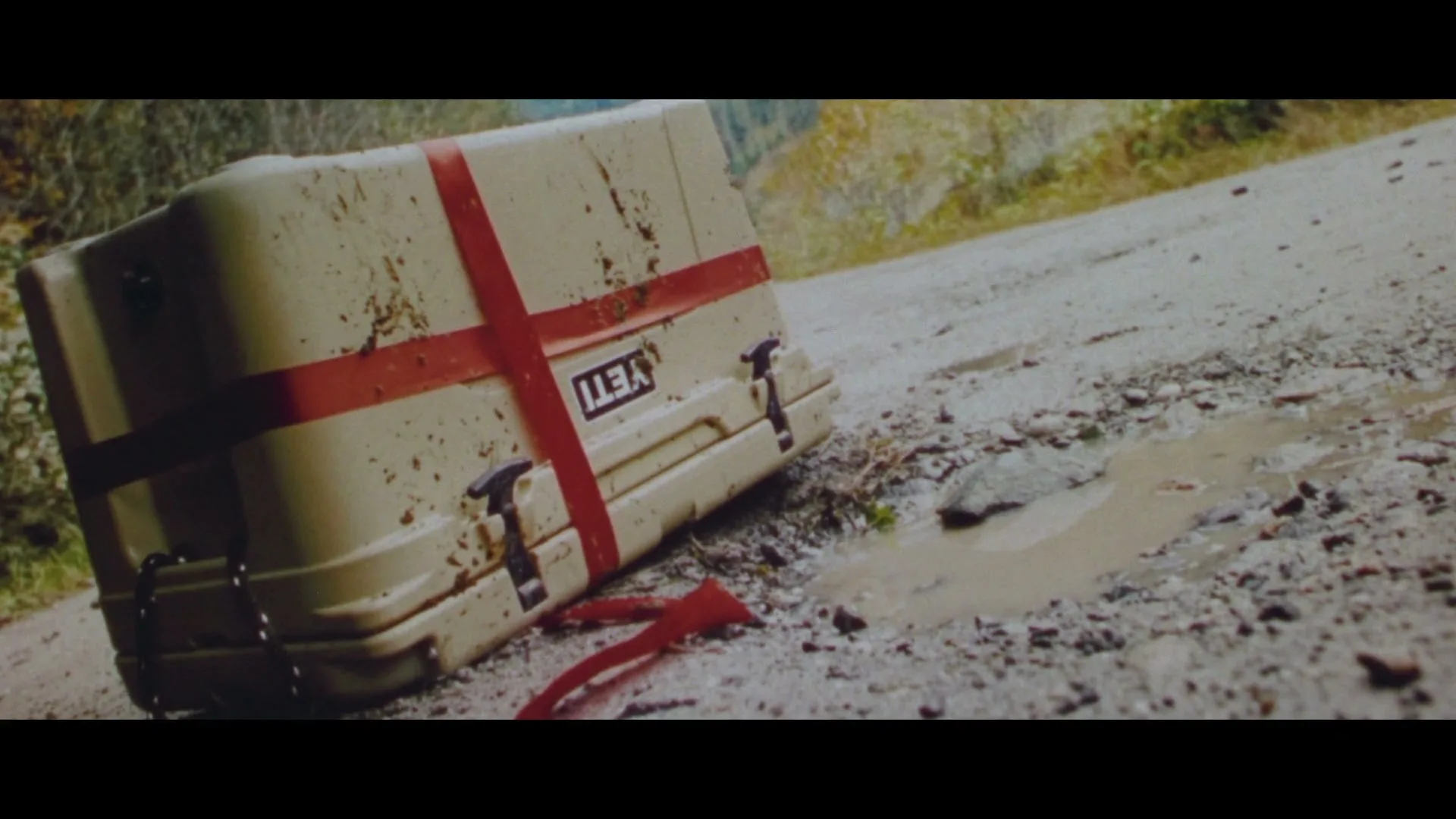 A YETI cooler, covered in mud and tied with a red ribbon, lies abandoned on a dirt road next to a puddle.