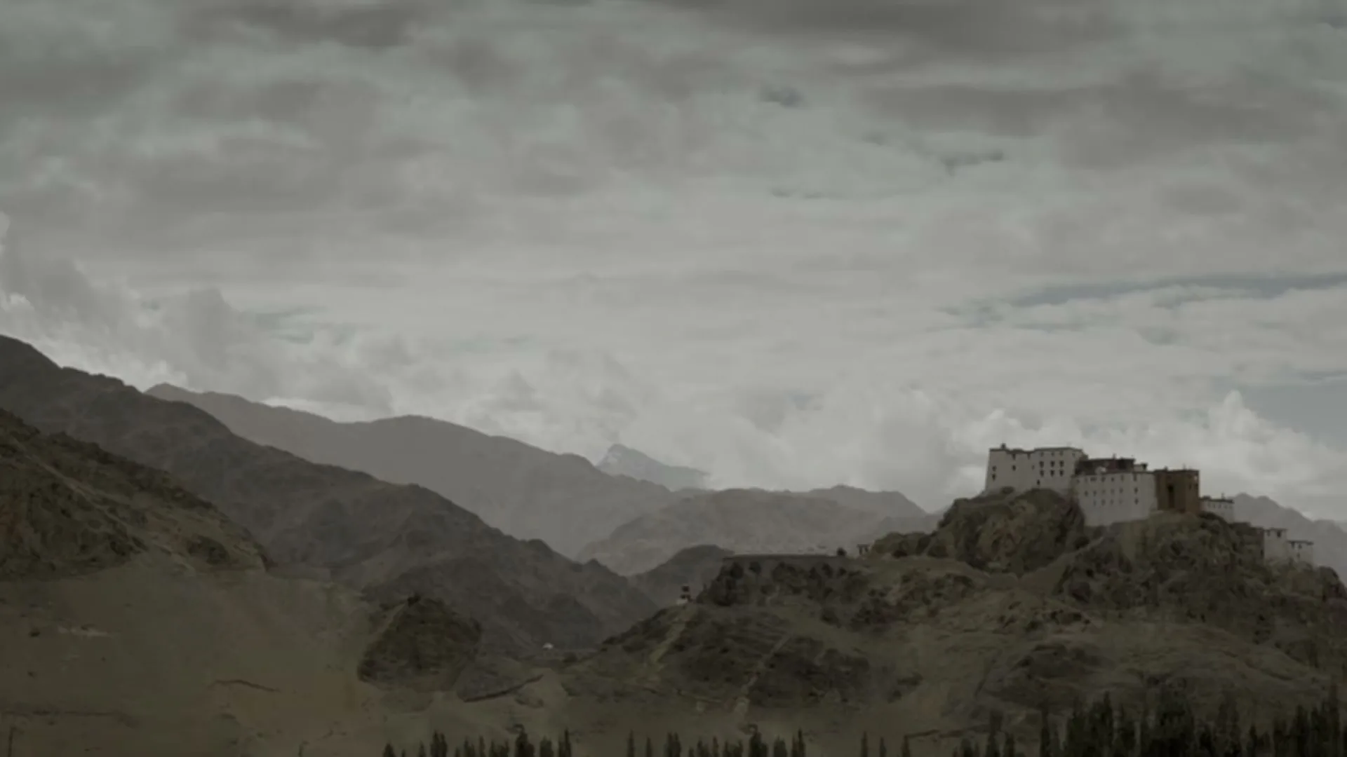 A wide shot shows a white monastery perched on a rocky hill, surrounded by vast, rugged mountains under a cloudy sky.