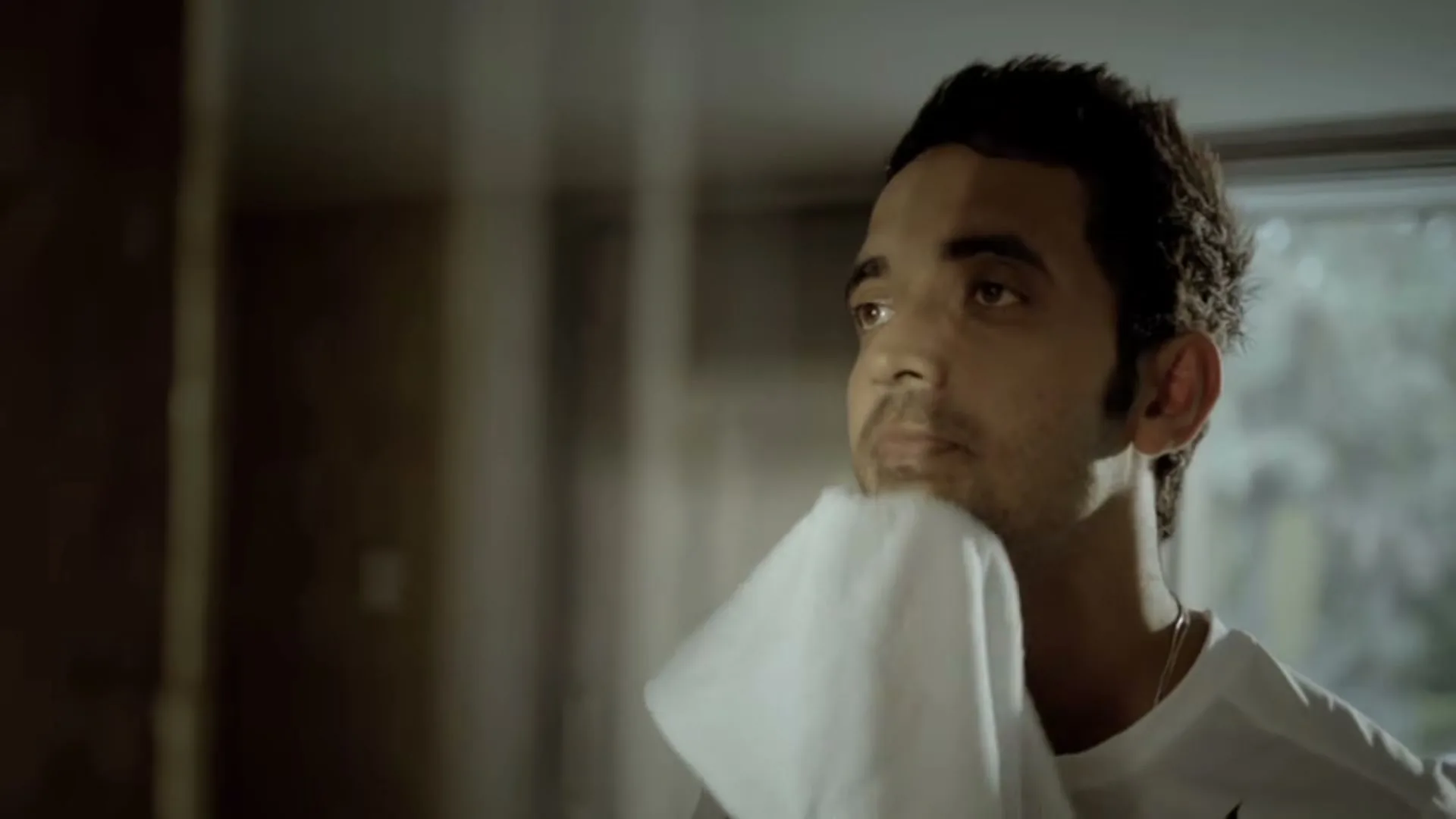 A young man with dark hair and a white t-shirt wipes his face with a white towel, looking up and to the right in an interior setting.
