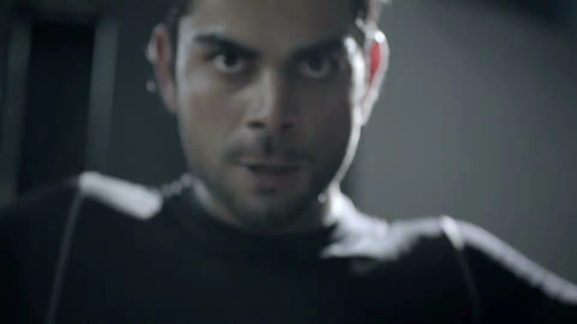 A determined male athlete with dark hair and a beard, wearing a black athletic shirt, looks intensely at the camera while exercising in a dimly lit gym.