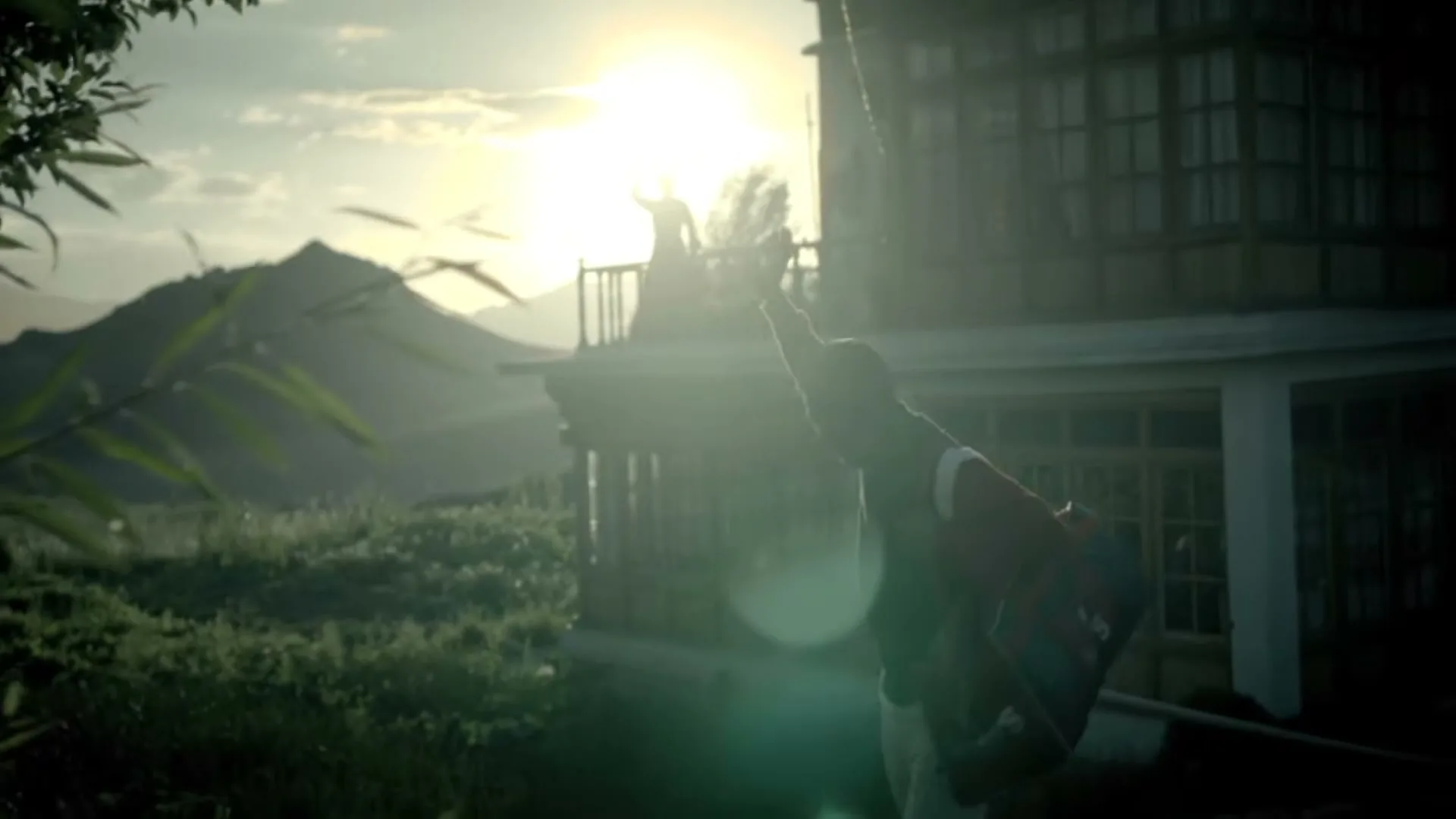 A young male athlete in a red and white jacket stretches his arm towards the bright sun, with mountains and a building in the background.