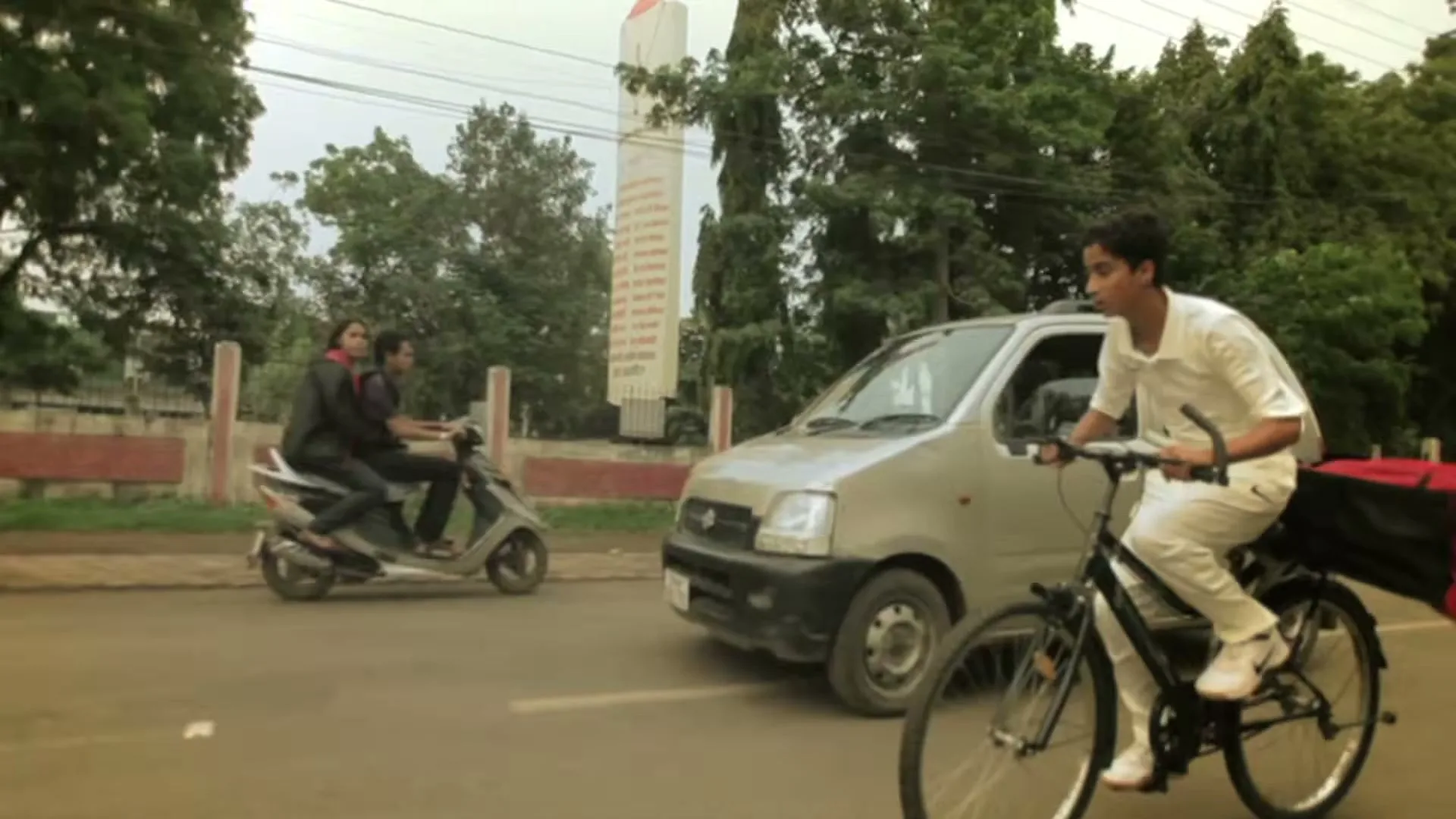 A young male cricketer in white uniform cycles on a road, passing a car and a scooter with two people, on a cloudy day.