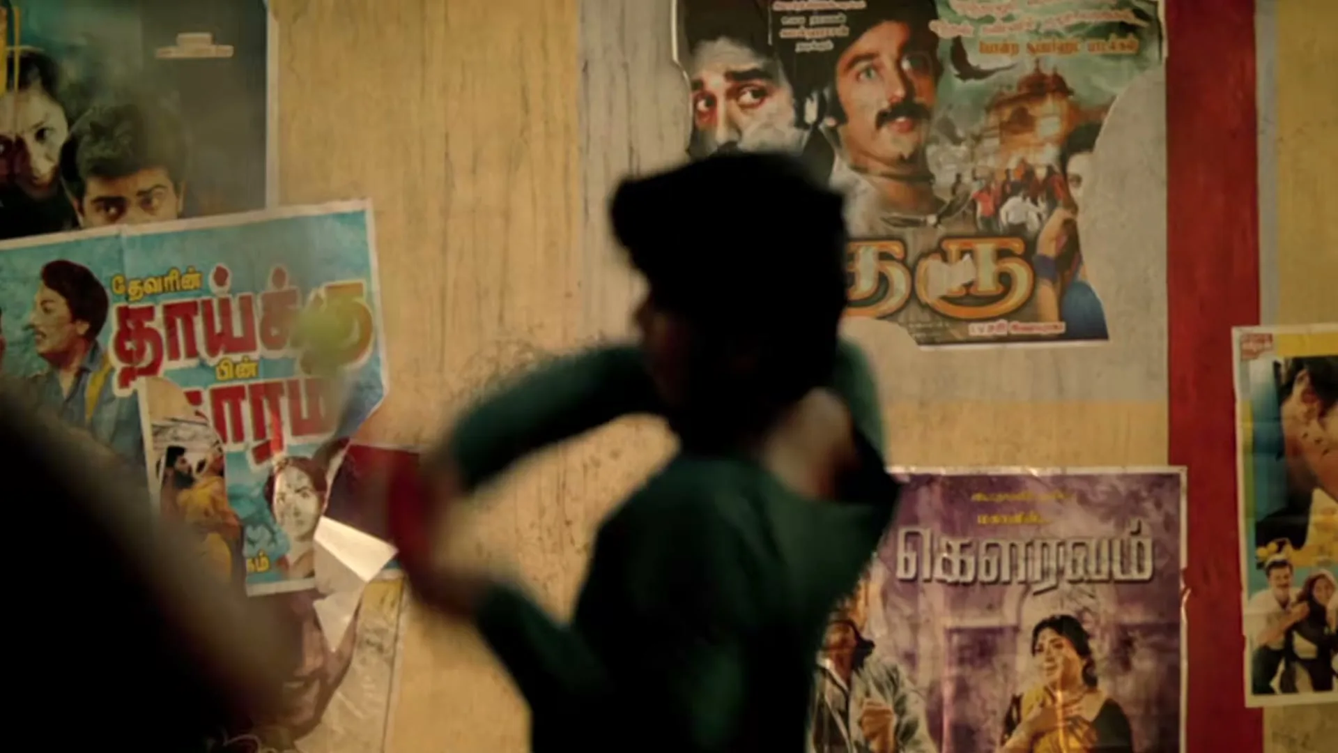 A young male athlete, blurred in the foreground, practices a tennis swing against a wall covered in colorful, torn movie posters in an urban setting.
