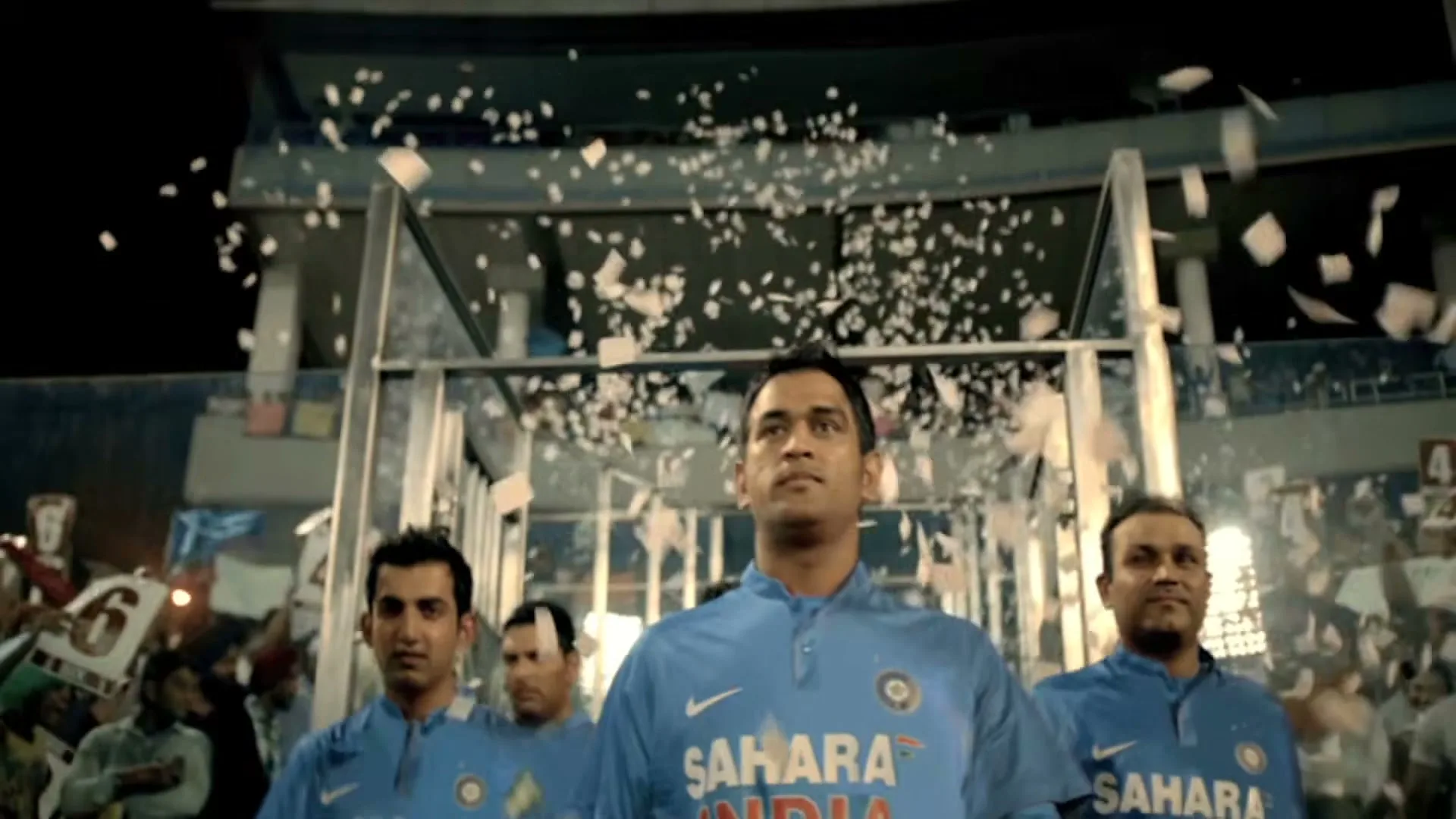 Indian cricket players, including MS Dhoni, walk onto a stadium field at night as confetti falls around them.