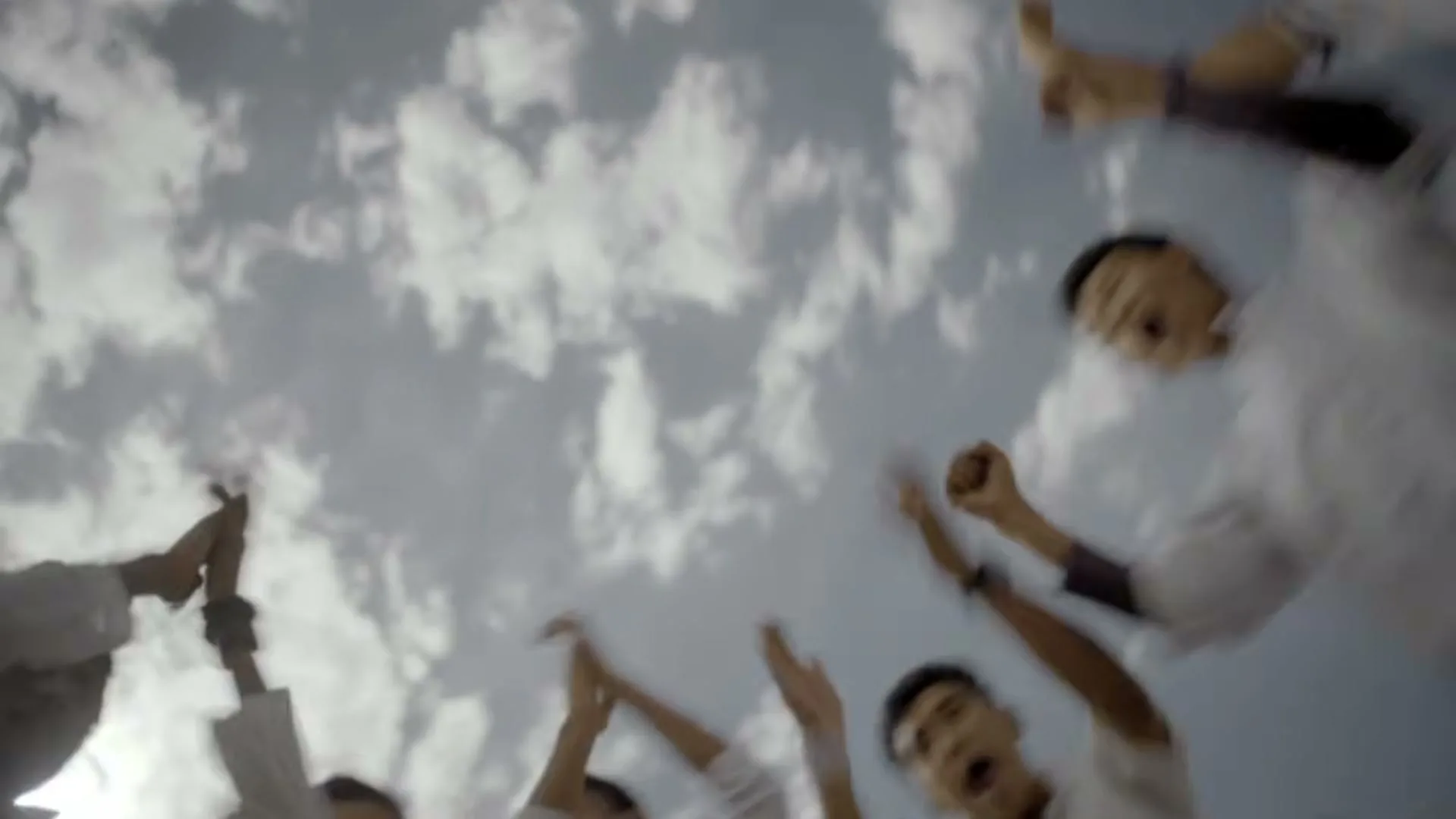 A group of young men in white shirts are seen from a low angle, cheering with their arms raised against a cloudy sky.