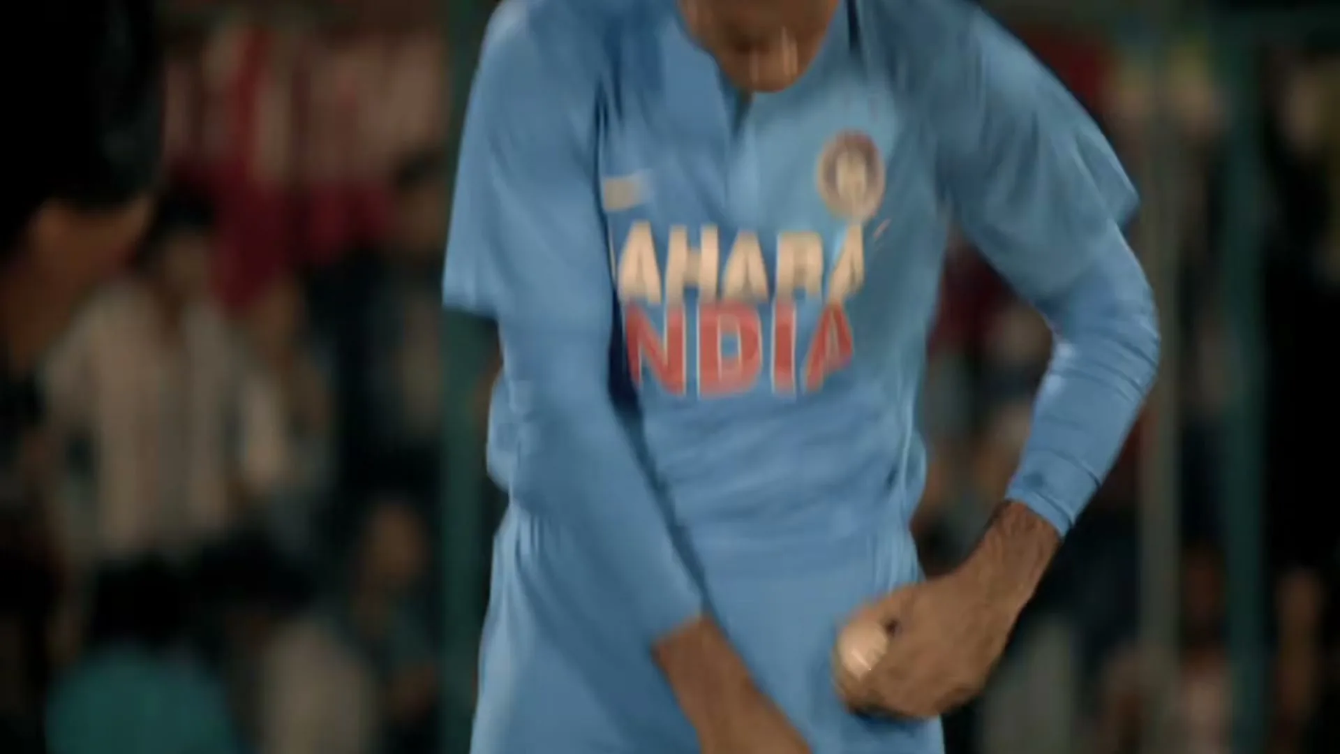A male cricket player in a blue uniform adjusts his protective gear, looking down with a focused expression on a sports field at night.